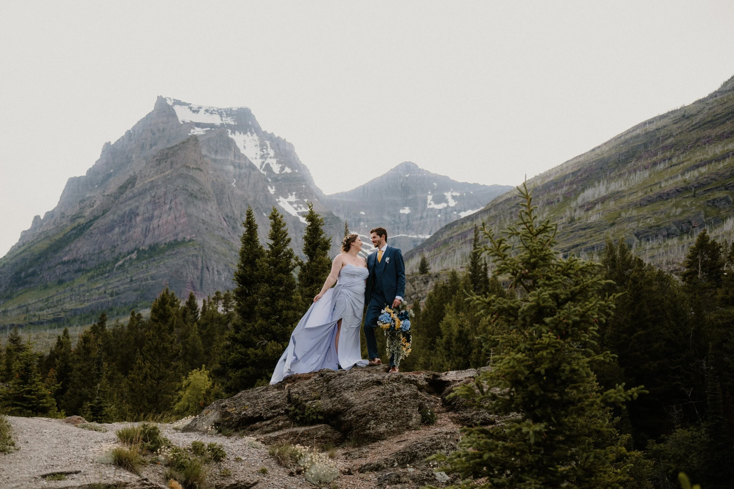 A bride and groom standing on a rocky ledge in a mountainous, forested area.