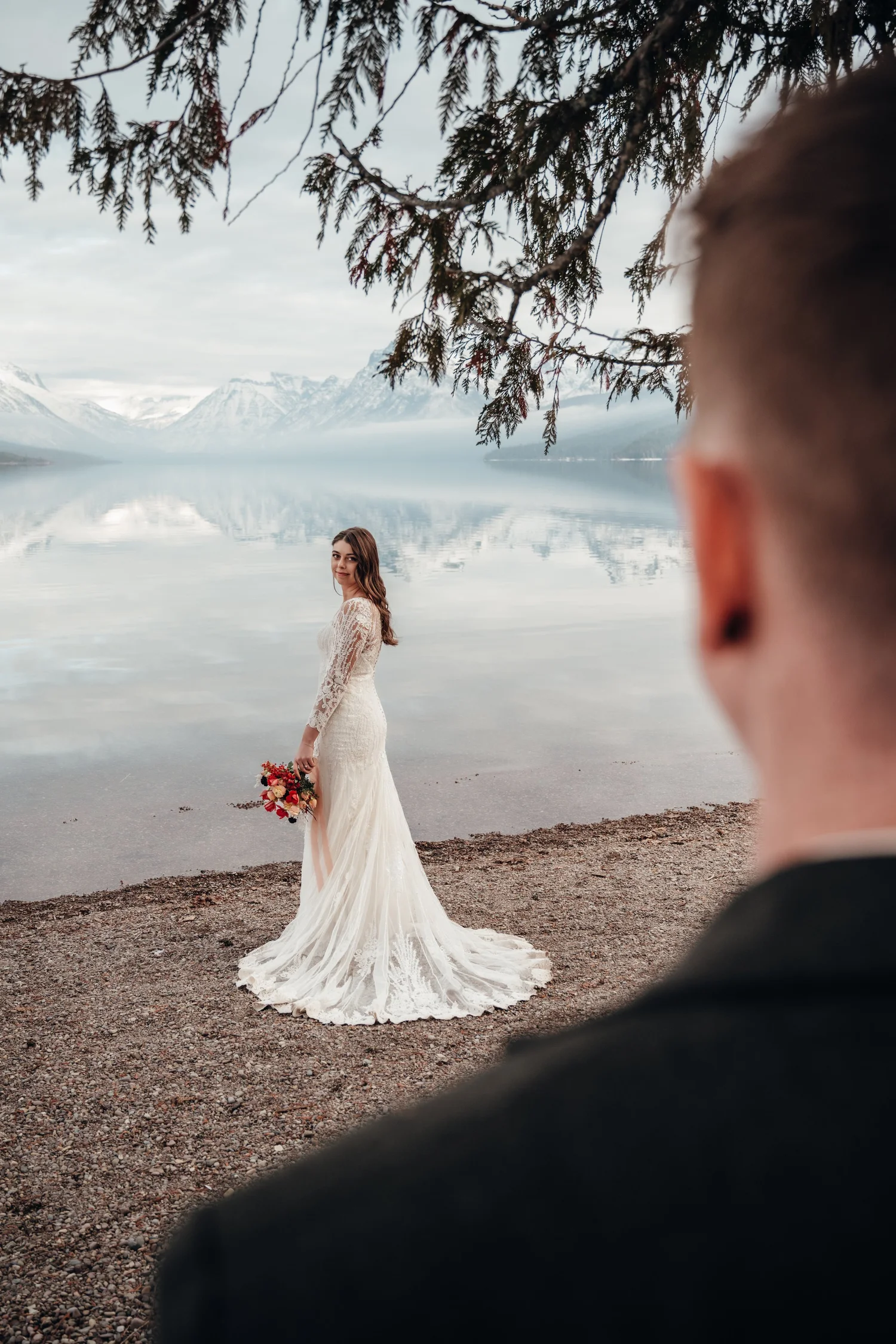 Bride in a lace gown holding her bouquet by Lake McDonald in December