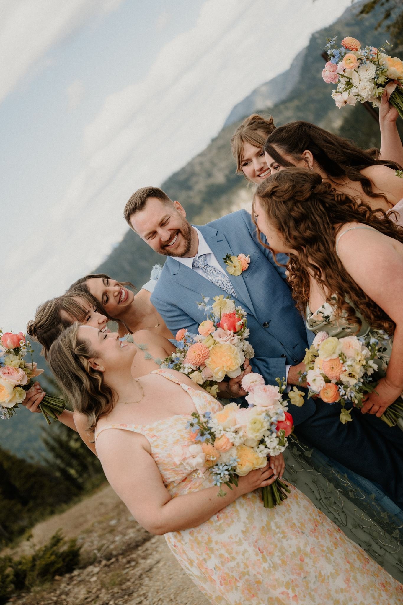 Group of six women and one man celebrate outdoors, holding colorful bouquets of flowers, dressed in pastel and floral attire, with mountains and cloudy sky in the background.