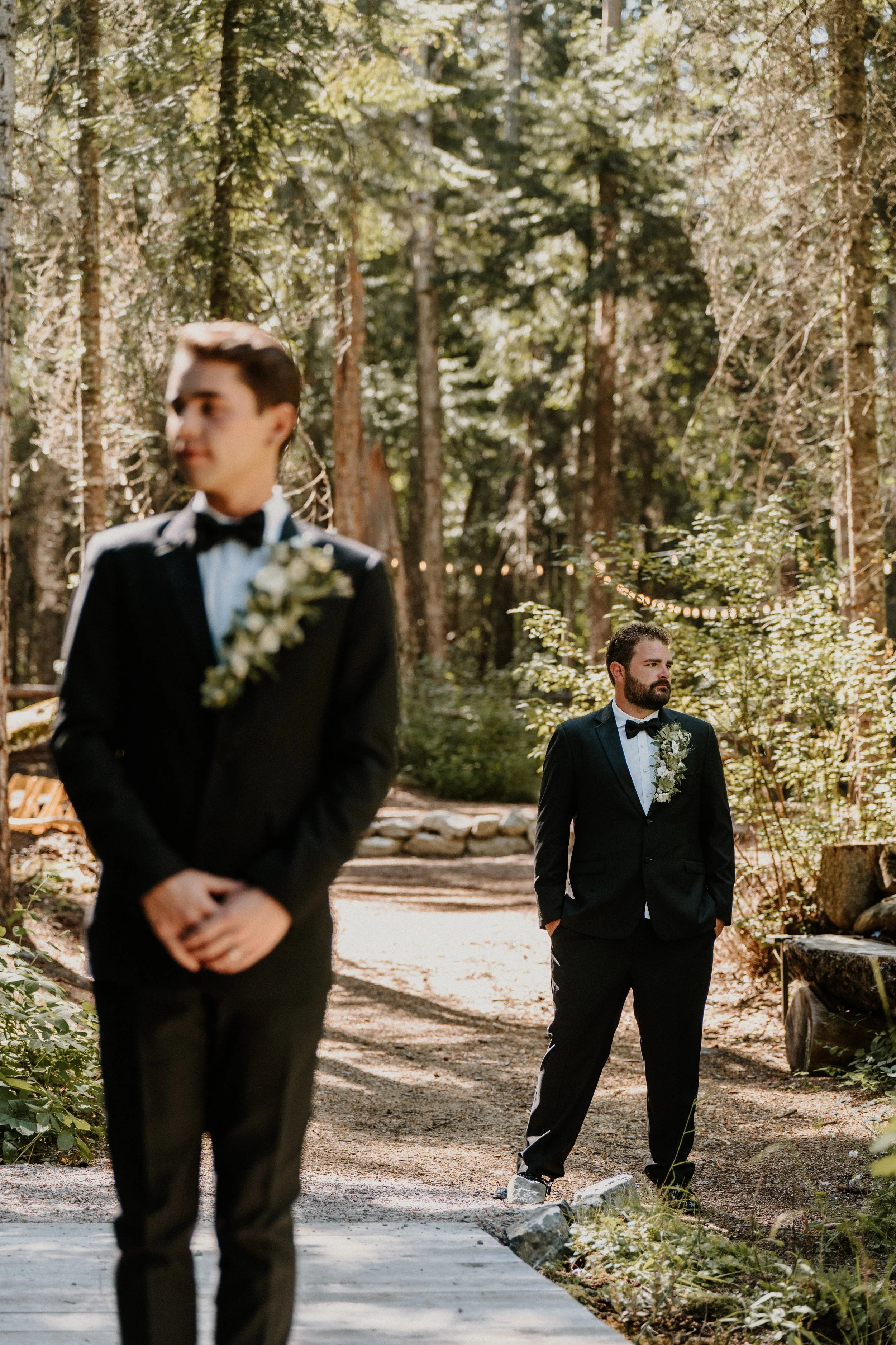 Two men in black tuxedos with floral boutonnieres are outdoors in a forested area, one in the foreground out of focus and the other in the background in focus.