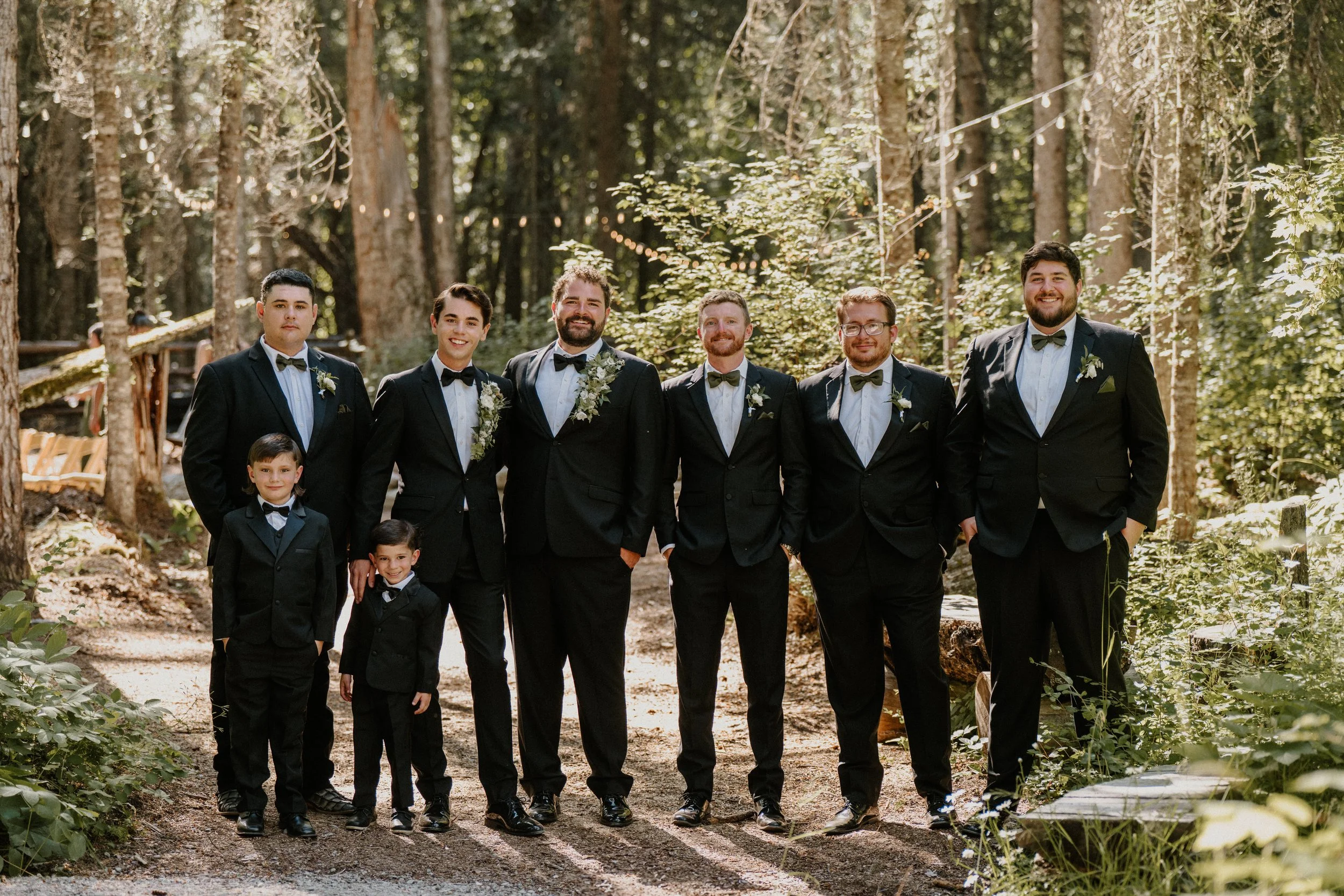Group of men and boys in black tuxedos with bow ties, standing outdoors in a wooded area, smiling for a photo.