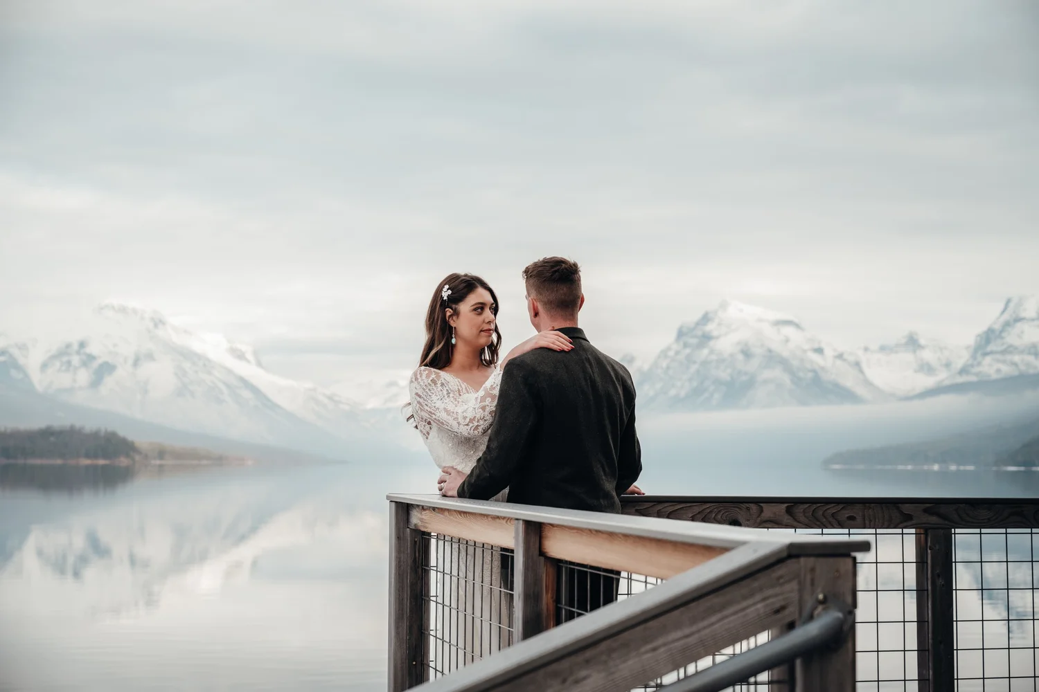 Couple leaning on the wooden dock railing at Apgar Village with snowy mountains behind Lake McDonald