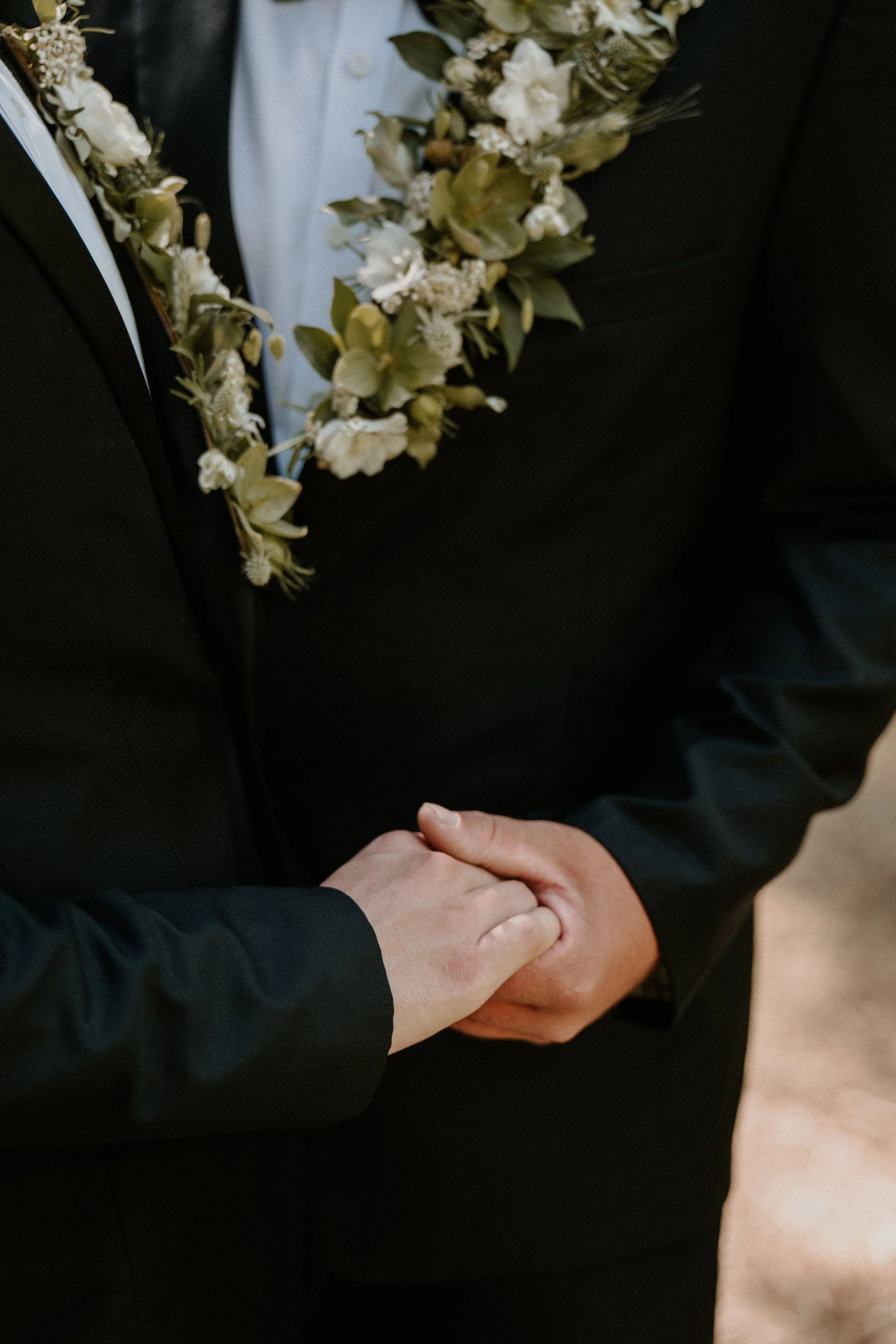 Close-up of a couple holding hands during a wedding or formal event, with the man wearing a black suit and a floral boutonnière.