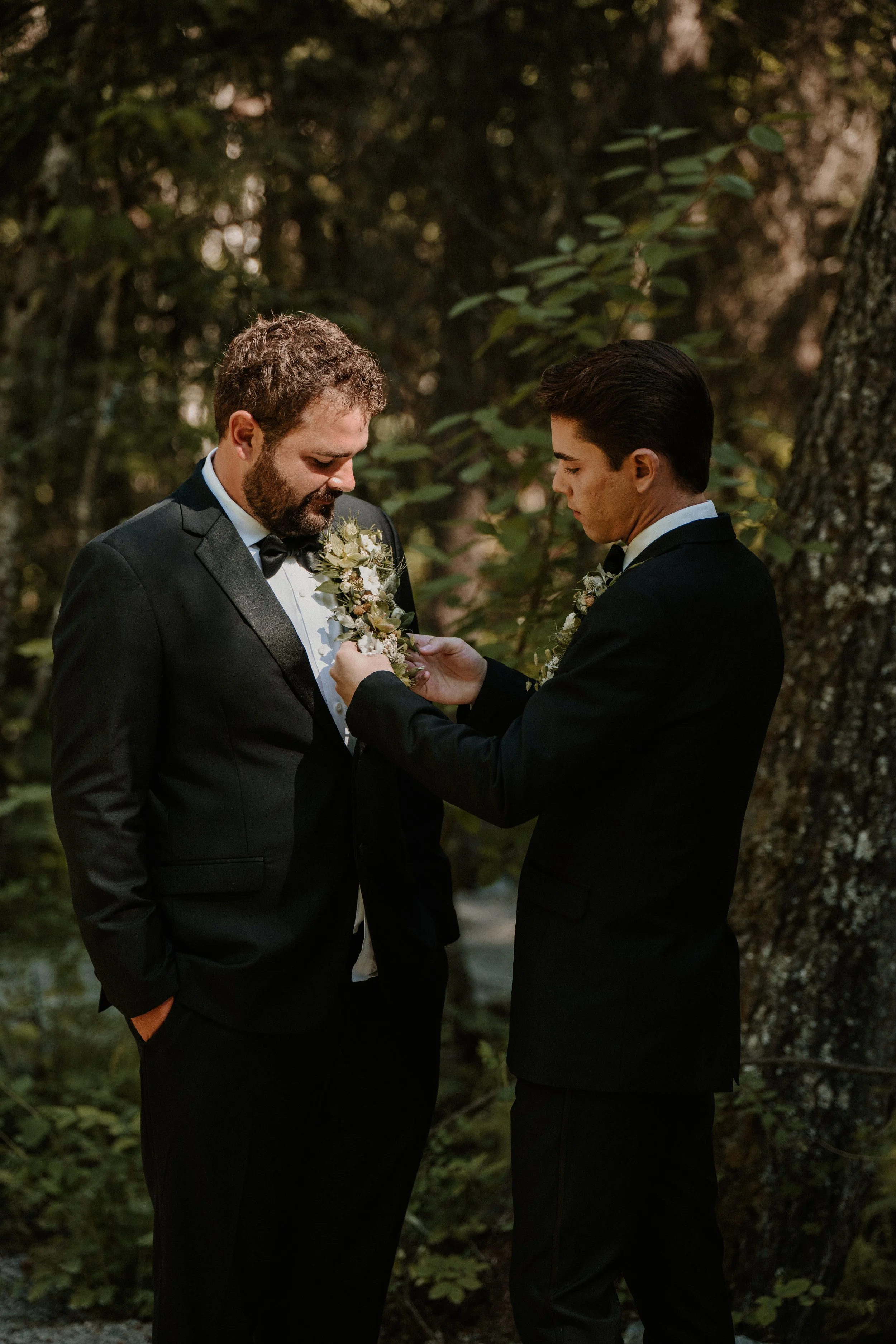 Two men in tuxedos standing outdoors with trees and foliage in the background. One man is pinning a boutonniere onto the other's lapel.