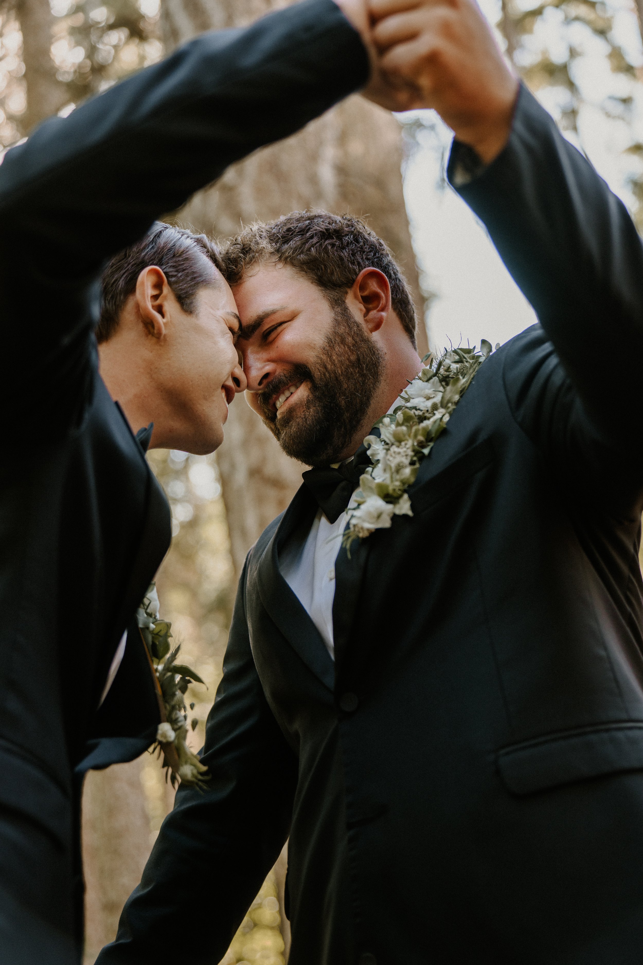 Two men in black tuxedos with white shirts and black bow ties, wearing floral boutonnieres, are embracing each other outdoors with their foreheads touching, smiling gently, in a forest setting with sunlight filtering through trees.