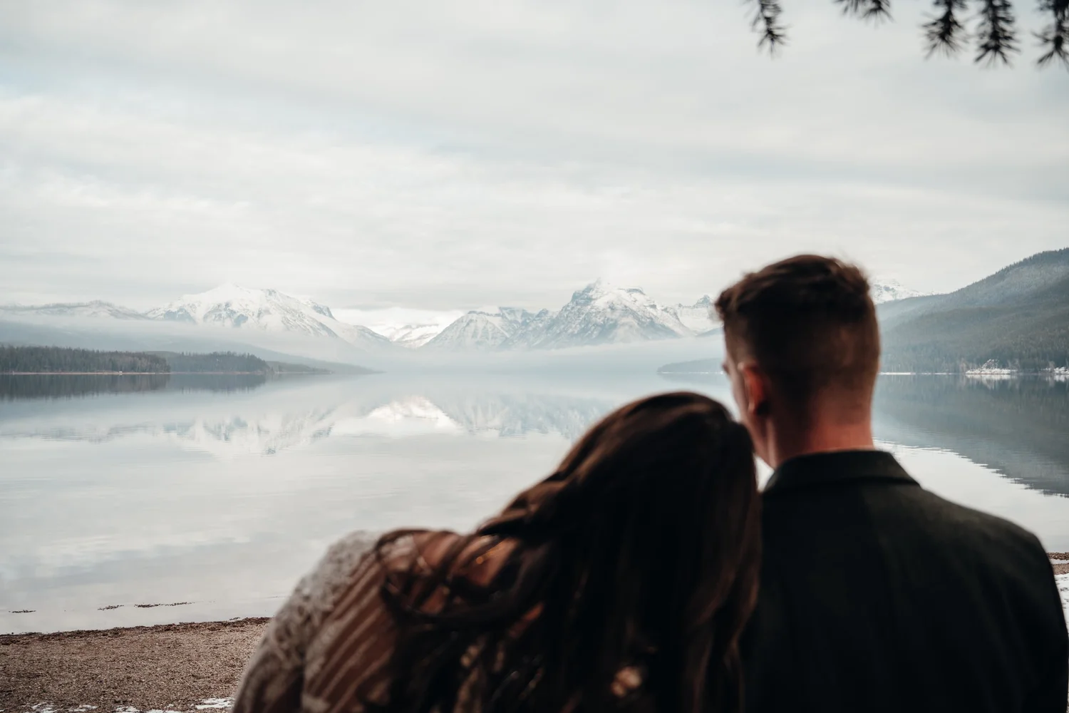 Couple looking out over a still Lake McDonald with snow-capped peaks reflected in the water