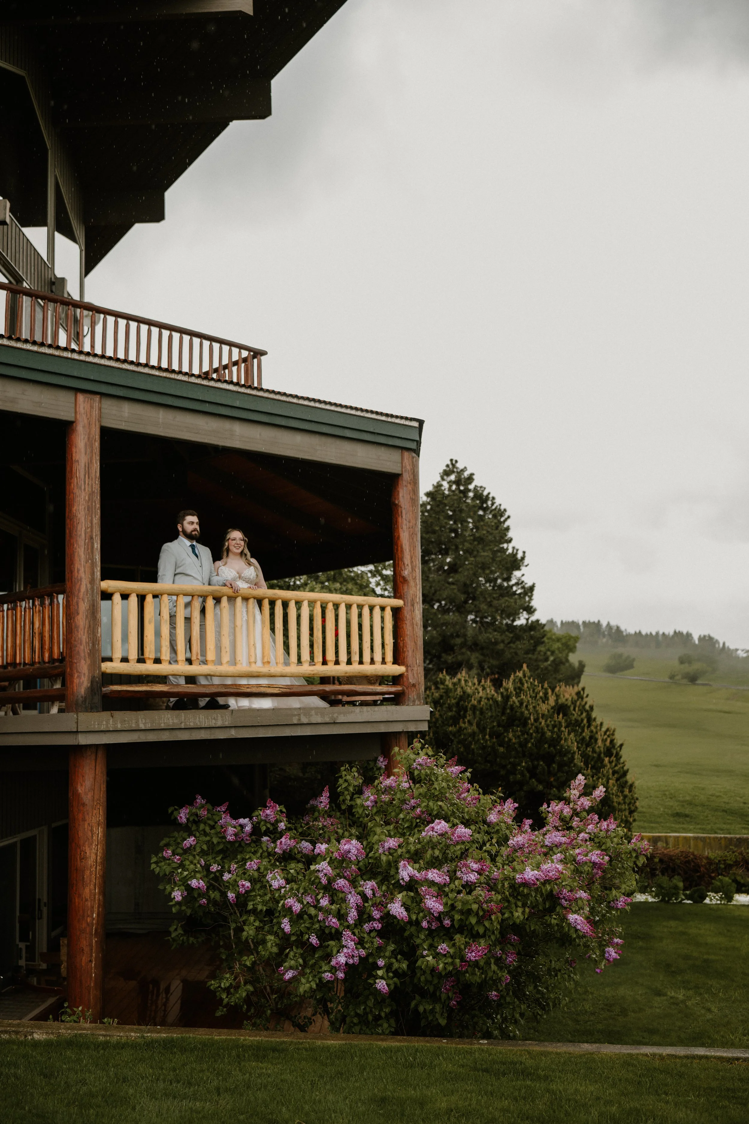 The couple on a wooden balcony with green hills and pink blossoms behind them.