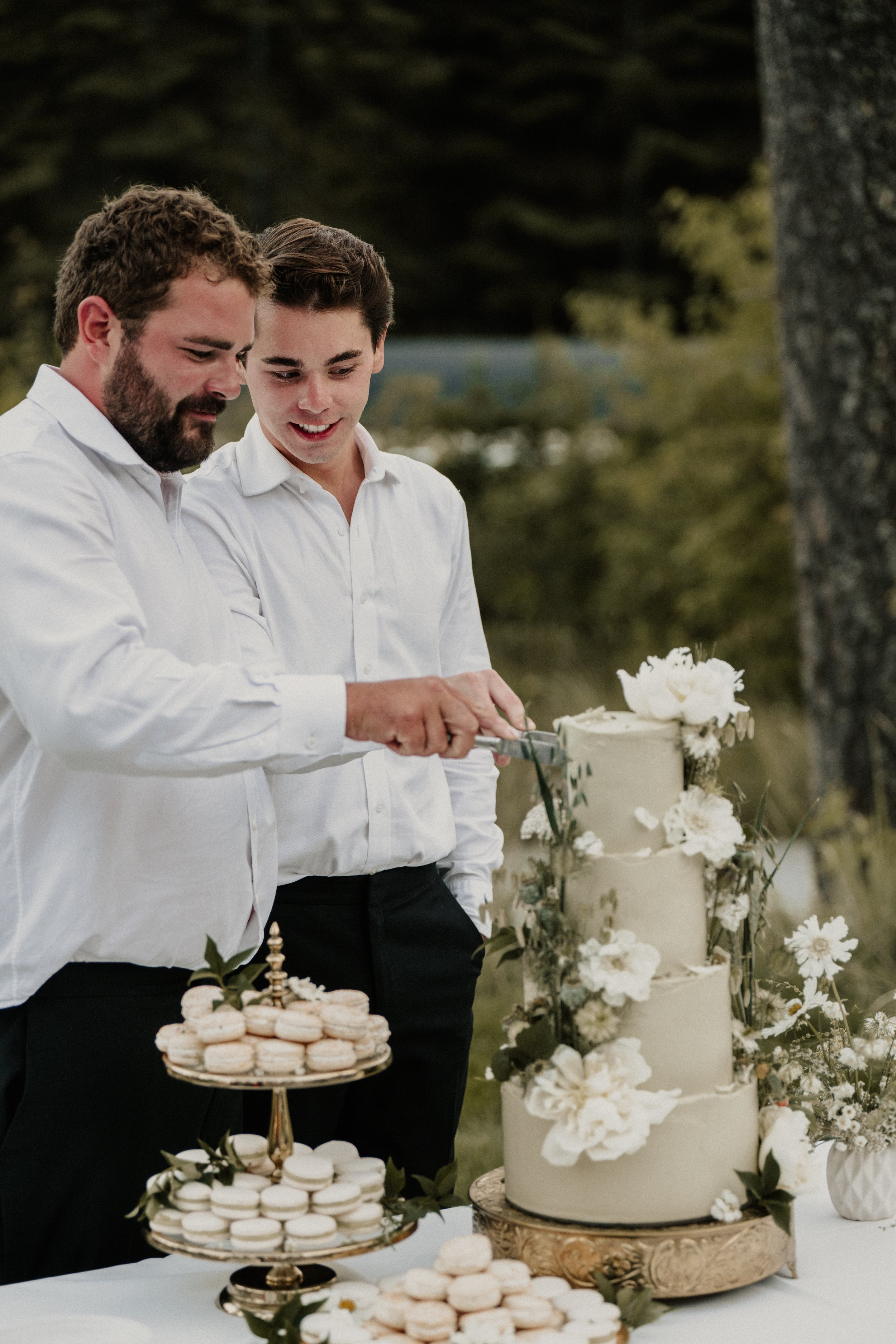 Two men in white shirts cutting into a four-tiered wedding cake decorated with white flowers, with macarons on a stand nearby, outdoors in a natural setting.