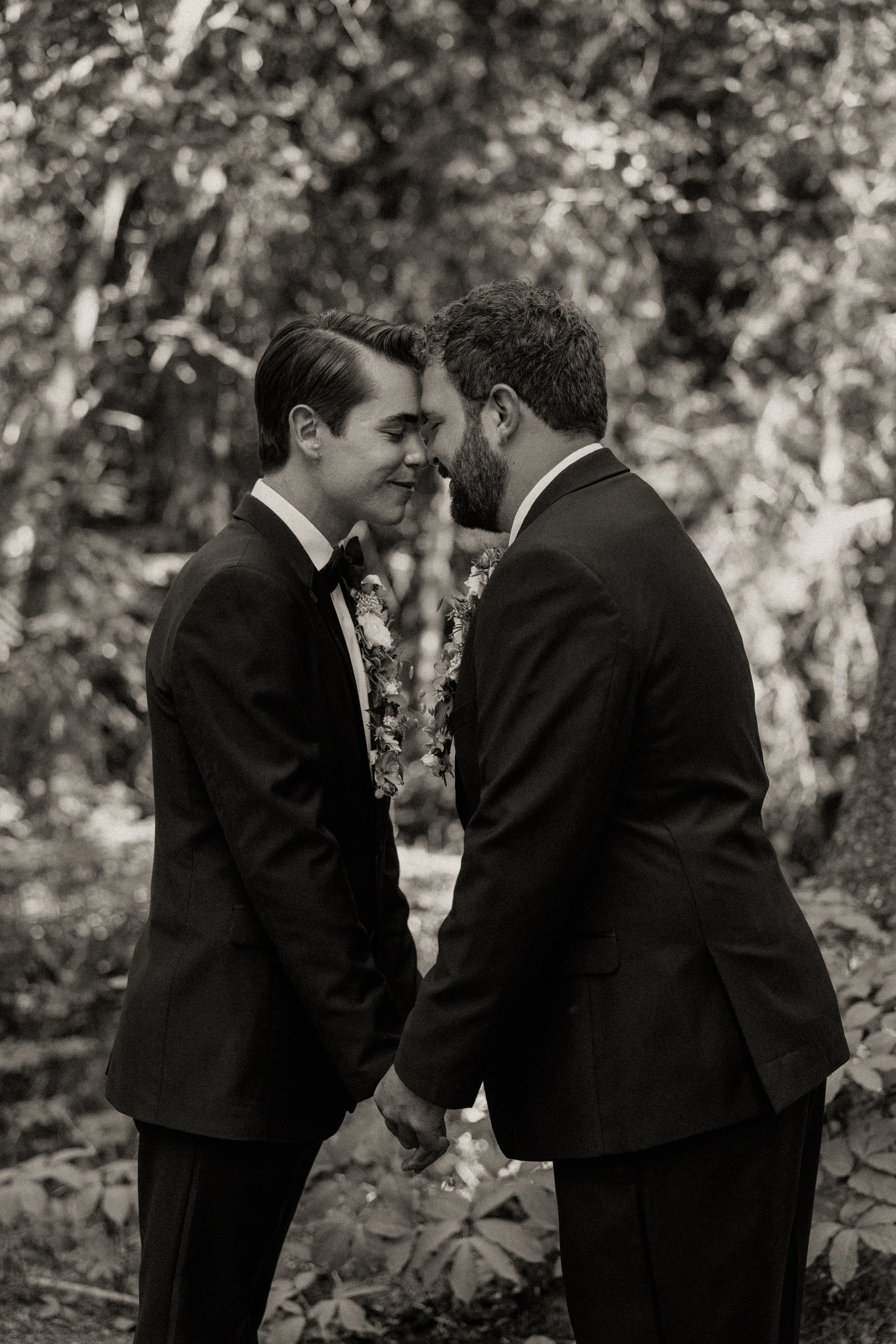 Two men in tuxedos holding hands and touching foreheads in a wedding or romantic ceremony outdoors, surrounded by trees.