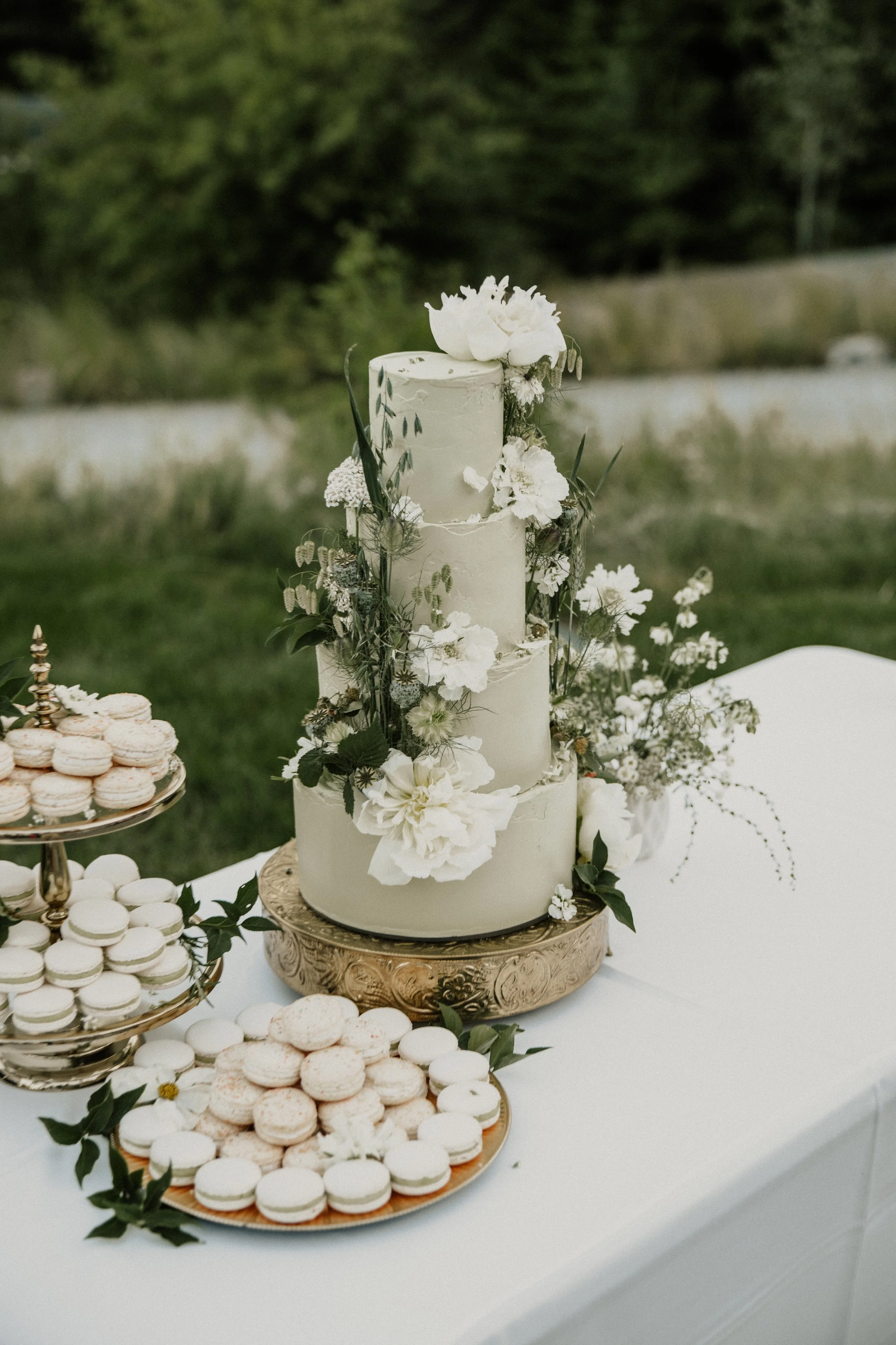 A three-tiered white wedding cake decorated with white flowers and greenery, surrounded by plates of macarons, set outdoors on a white table.