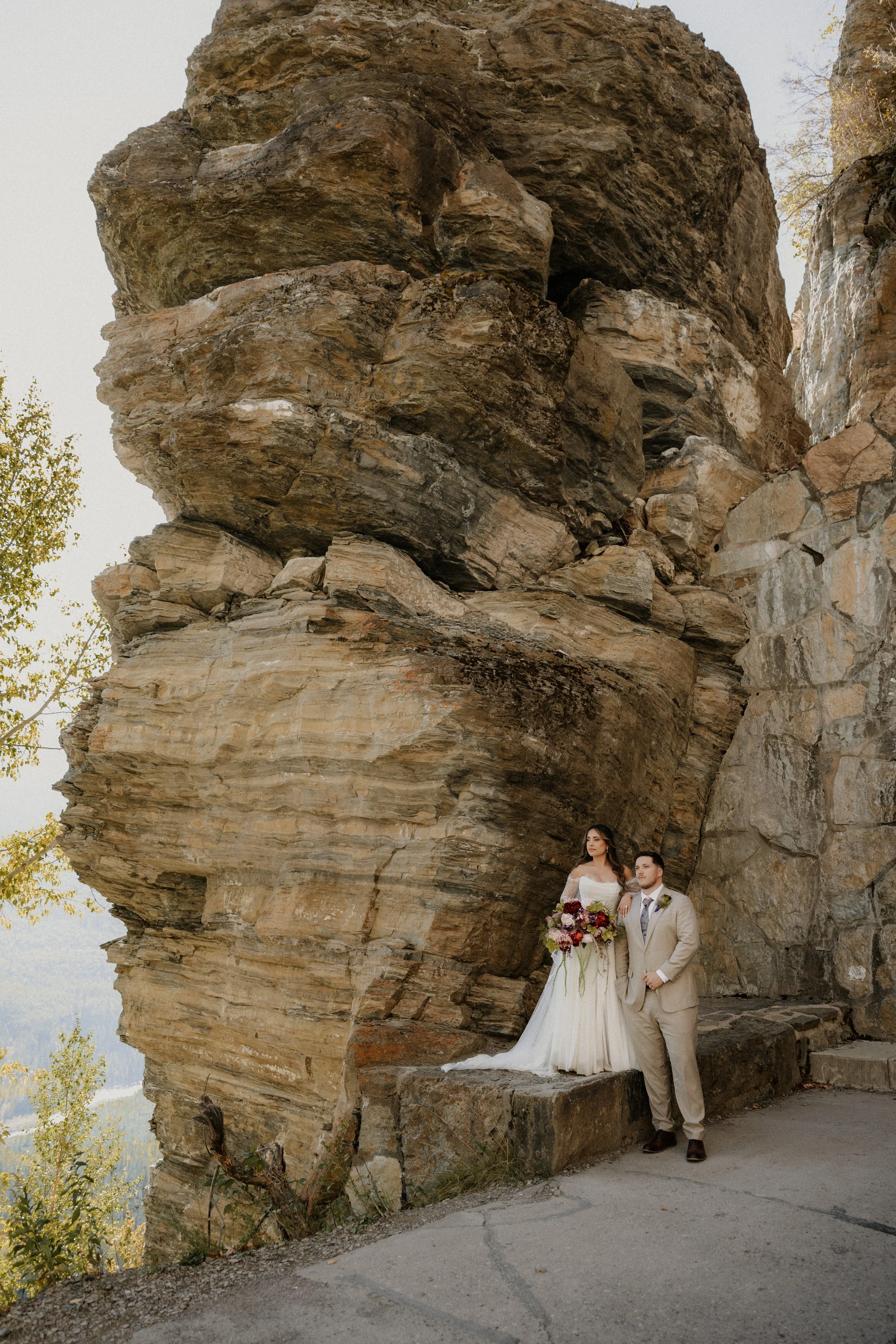 A bride and groom standing in front of a large rock formation during their wedding. The bride wears a white wedding gown and holds a bouquet of flowers. The groom wears a beige suit and stands close to the bride, both looking towards the camera.