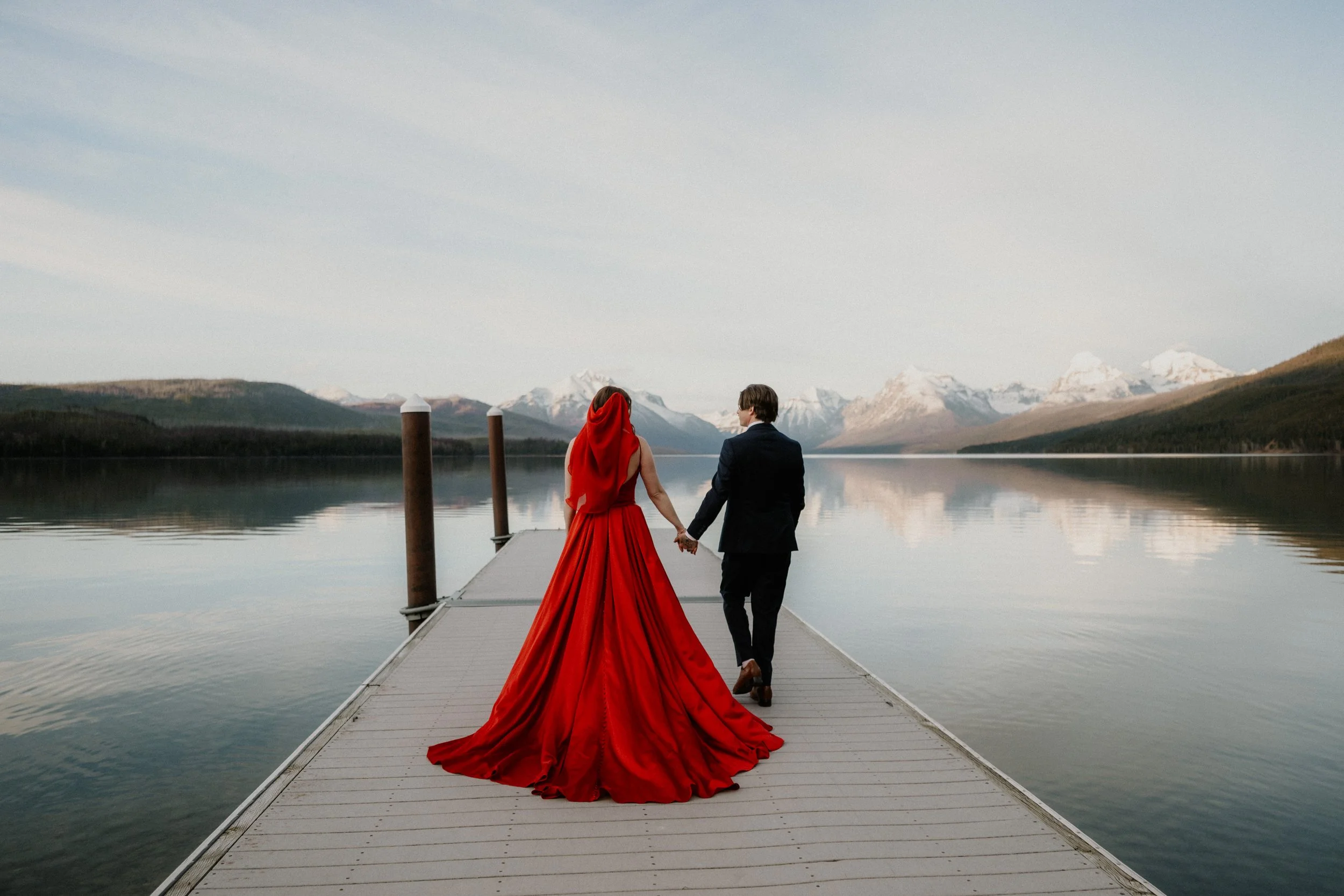 Couple walking a lake dock, red dress and hood, snow-capped mountains