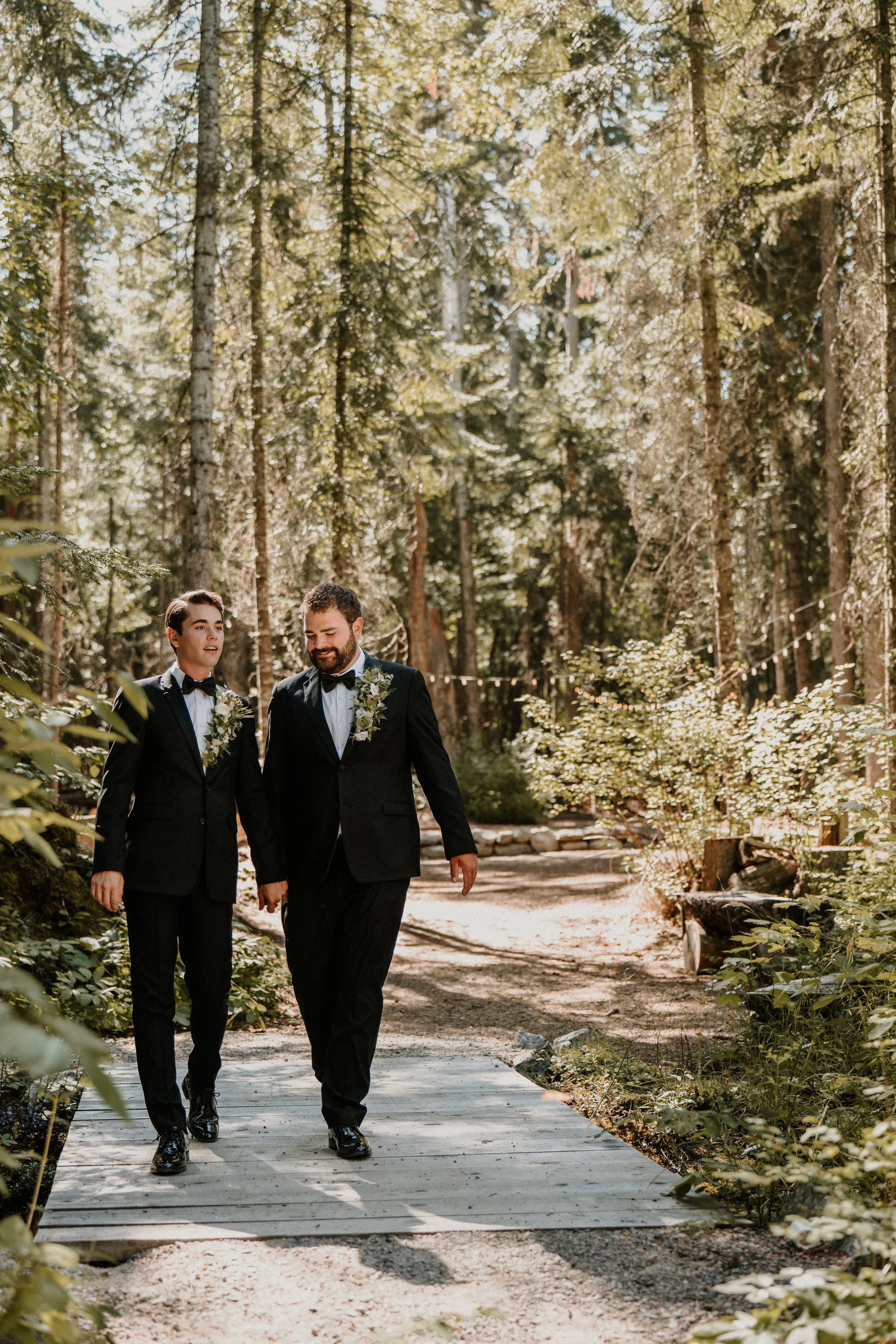Two men in tuxedos walk hand in hand on a wooden path through a forest, decorated with flowers on their jackets, during daytime.