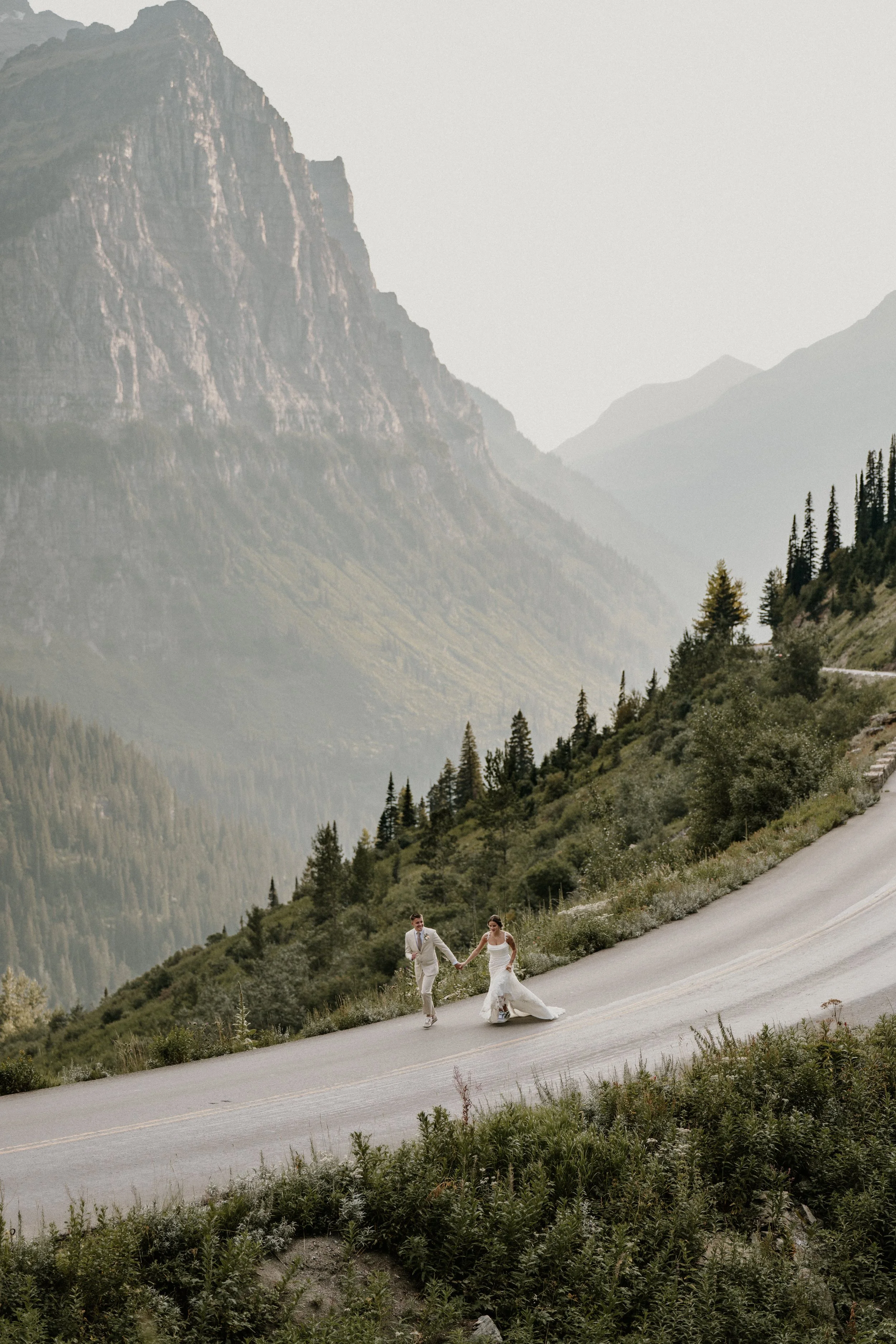 A bride and groom holding hands and walking on a mountain road surrounded by lush greenery and tall evergreen trees, with mountains in the background.