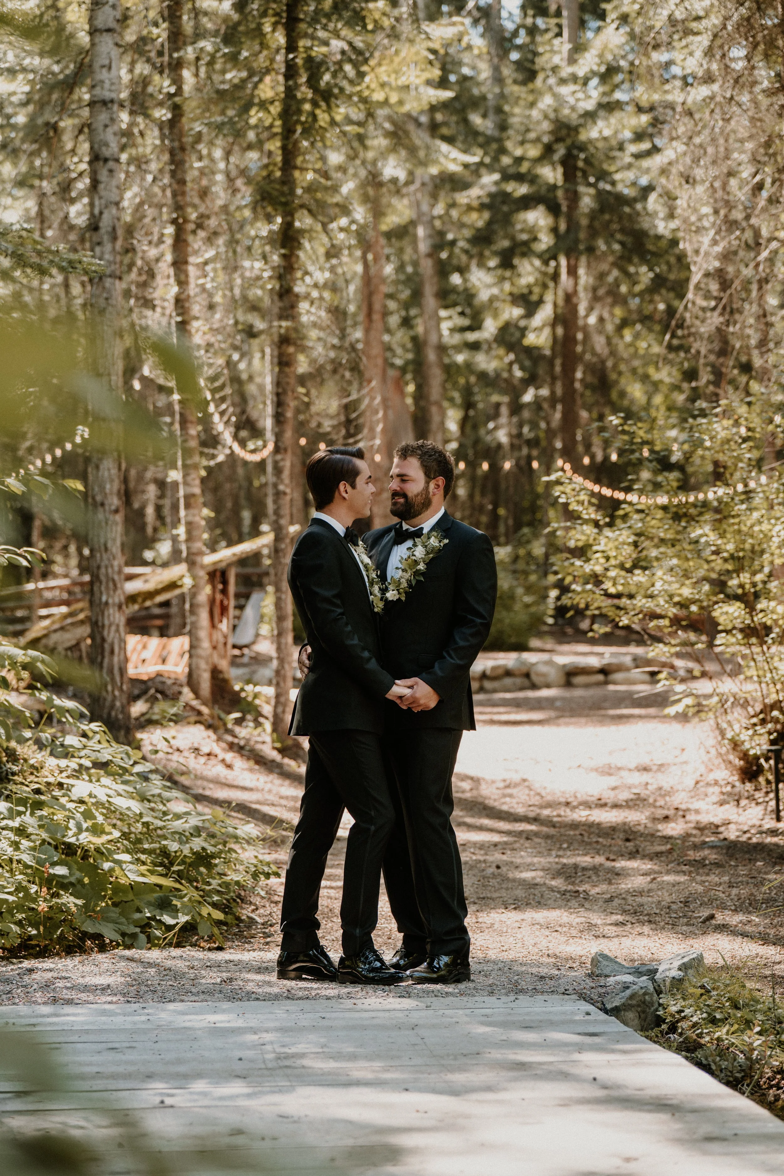 A same-sex couple dressed in black tuxedos holding hands and smiling at each other in a forest setting during a wedding ceremony, with string lights in the background.