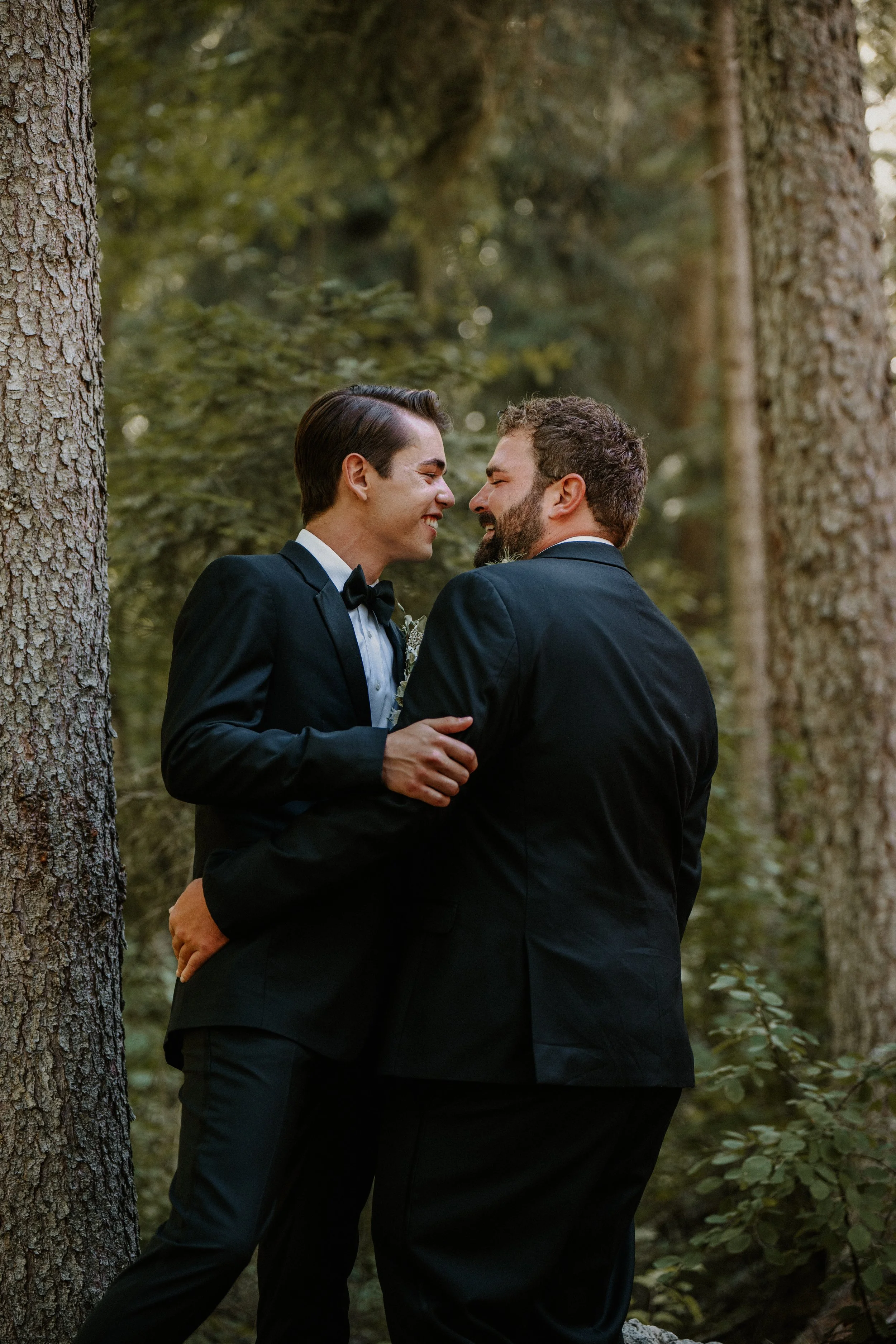 A same-sex couple in formal attire sharing a kiss in a forest.