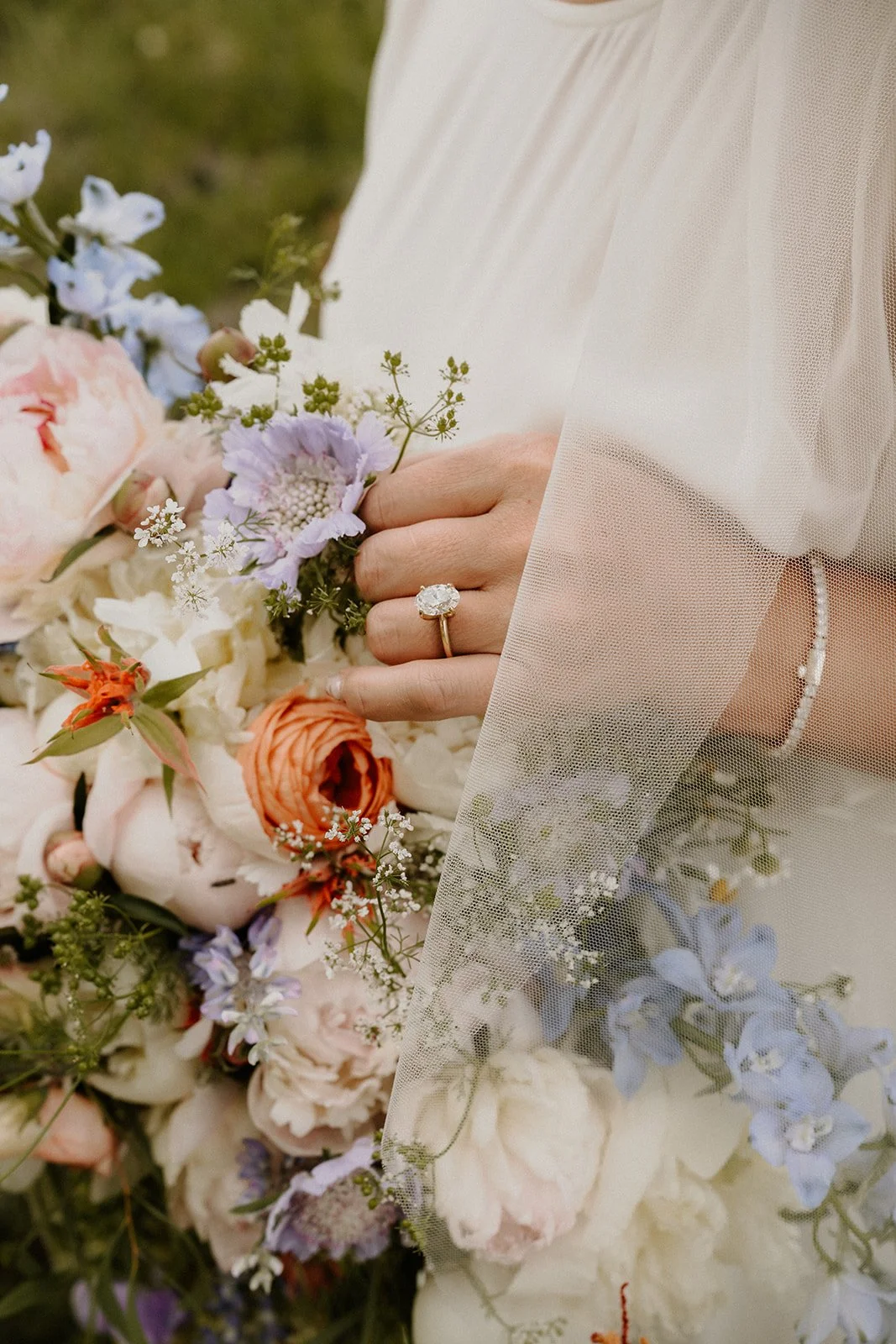 Close-up of a woman's hand wearing a diamond ring, holding a bouquet of pastel-colored flowers, with sheer fabric overlaying part of the image.