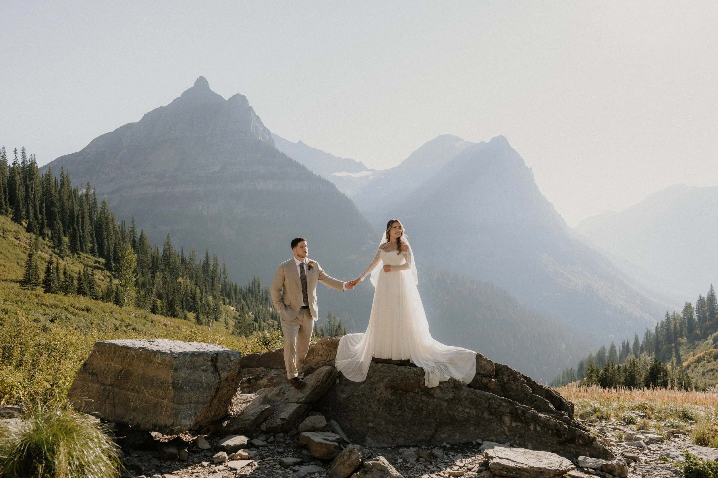 A wedding couple standing on a rocky ledge in a mountainous landscape with tall pine trees and high peaks in the background. The bride is wearing a white wedding dress and the groom in a beige suit holding hands.
