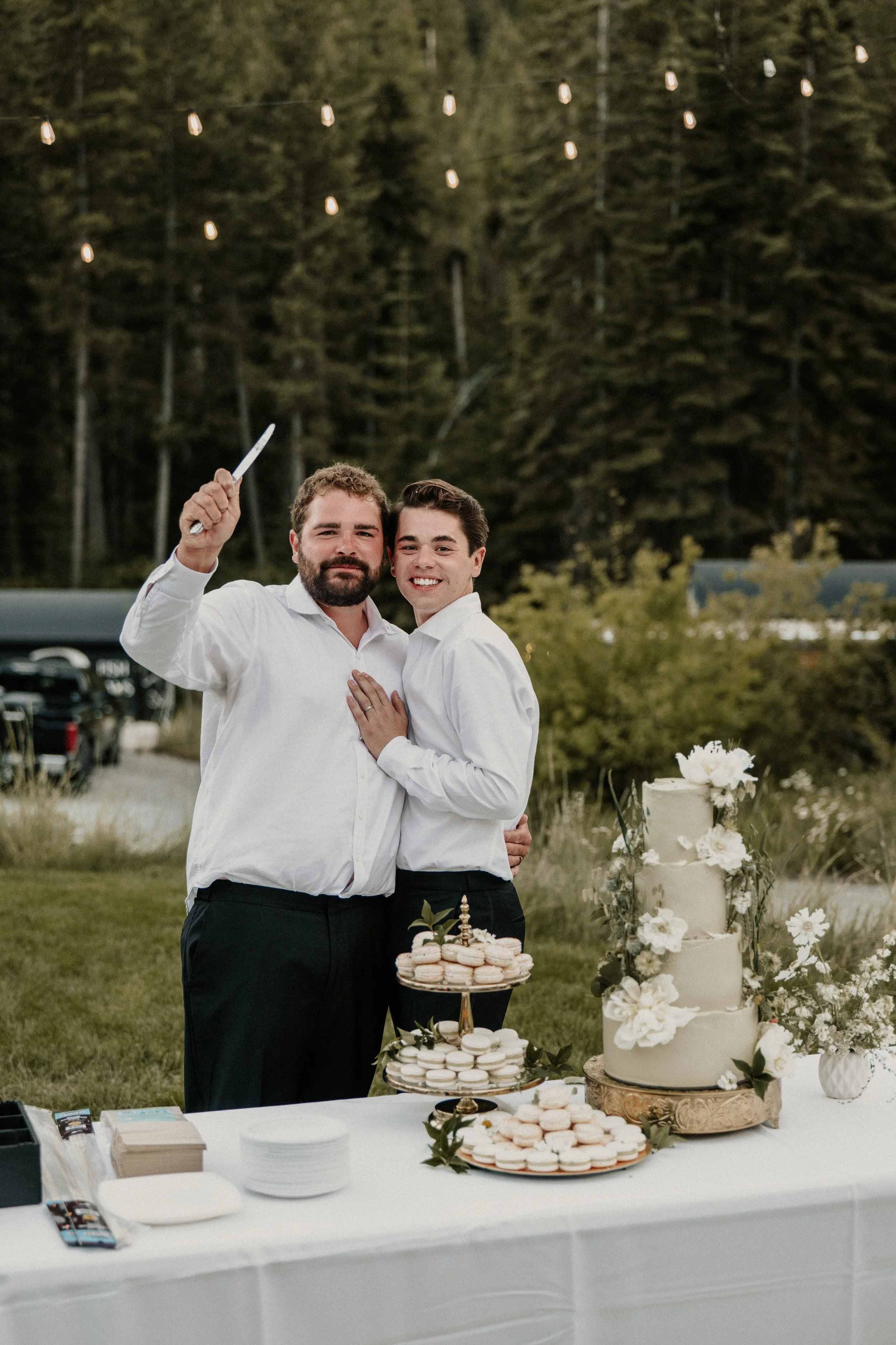 Two men in white shirts smiling and embracing at an outdoor wedding reception, with a tiered wedding cake decorated with flowers and macarons in front of them.