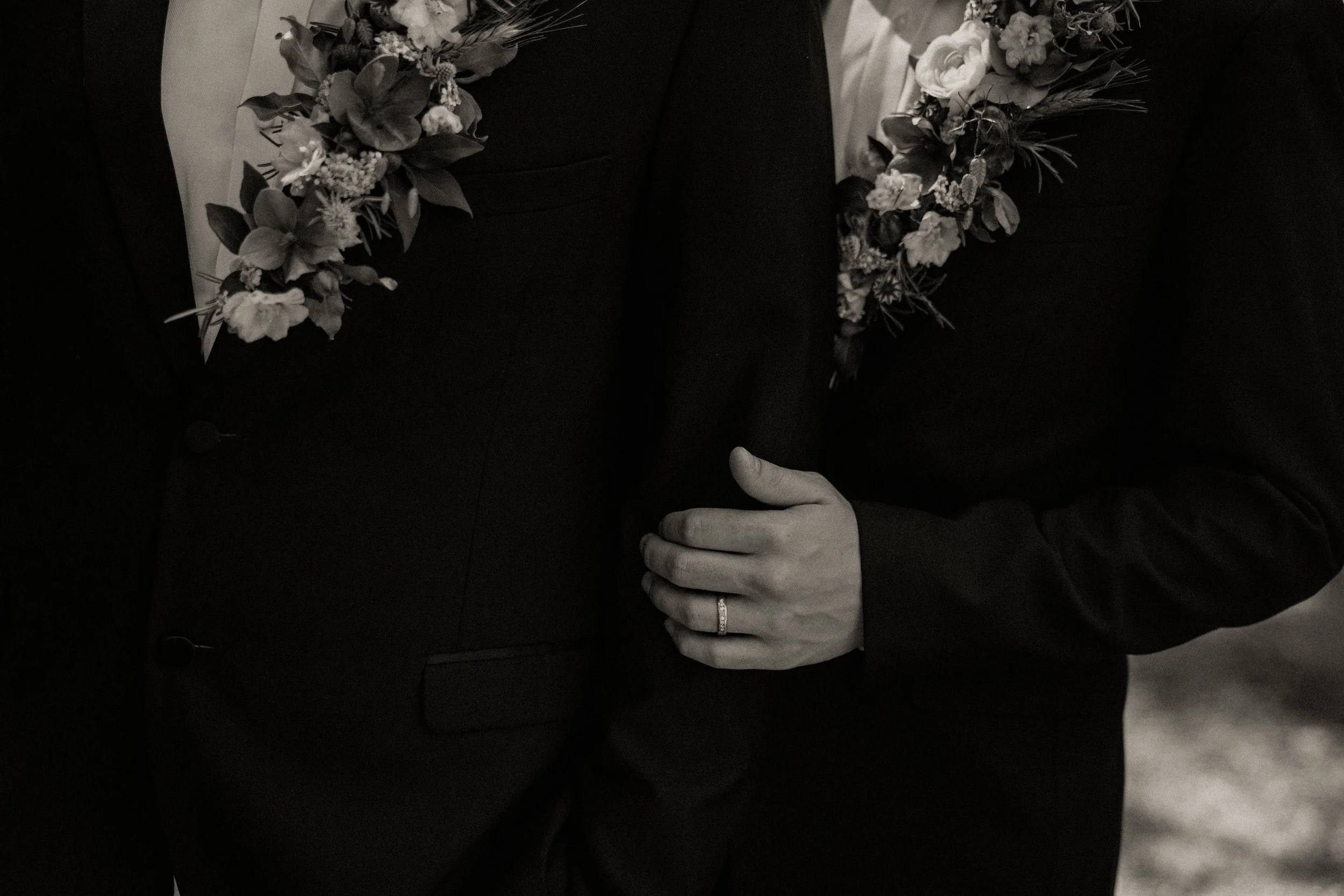 Close-up of a wedding couple in formal attire backstage or at an event, with focus on the woman's hand showing a wedding ring, and floral leis draped around the men's necks.