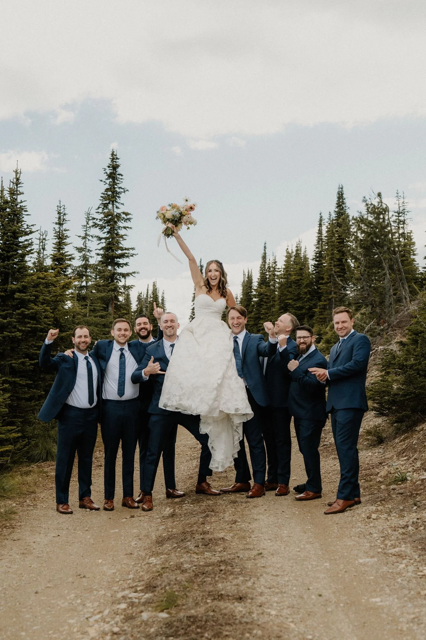 A bride in a white wedding dress on the shoulders of a group of men in suits, holding a bouquet up in the air, outdoors in a forested area with trees and cloudy sky.
