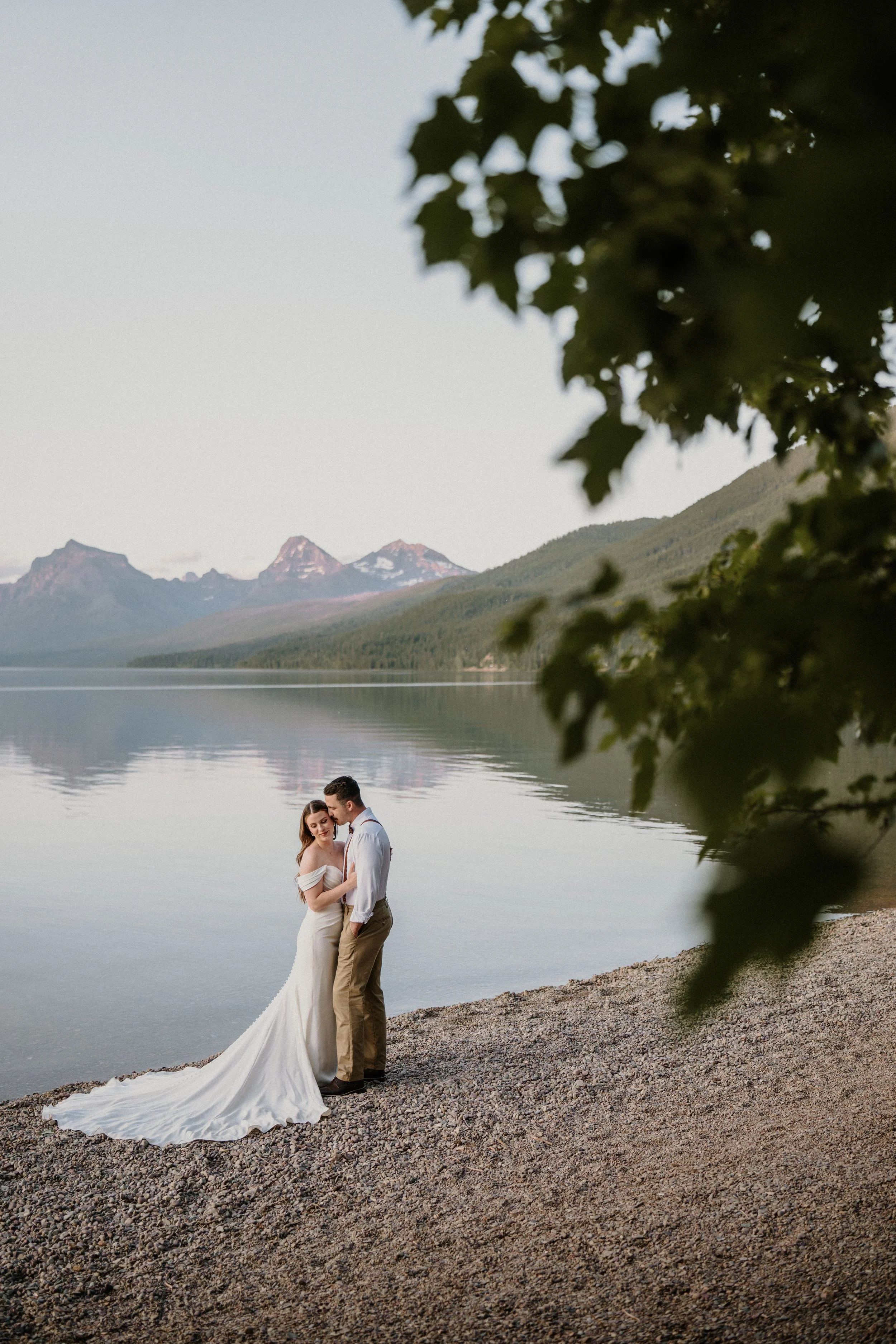 A couple stands on a rocky shore by a calm lake with mountains in the background, surrounded by trees.
