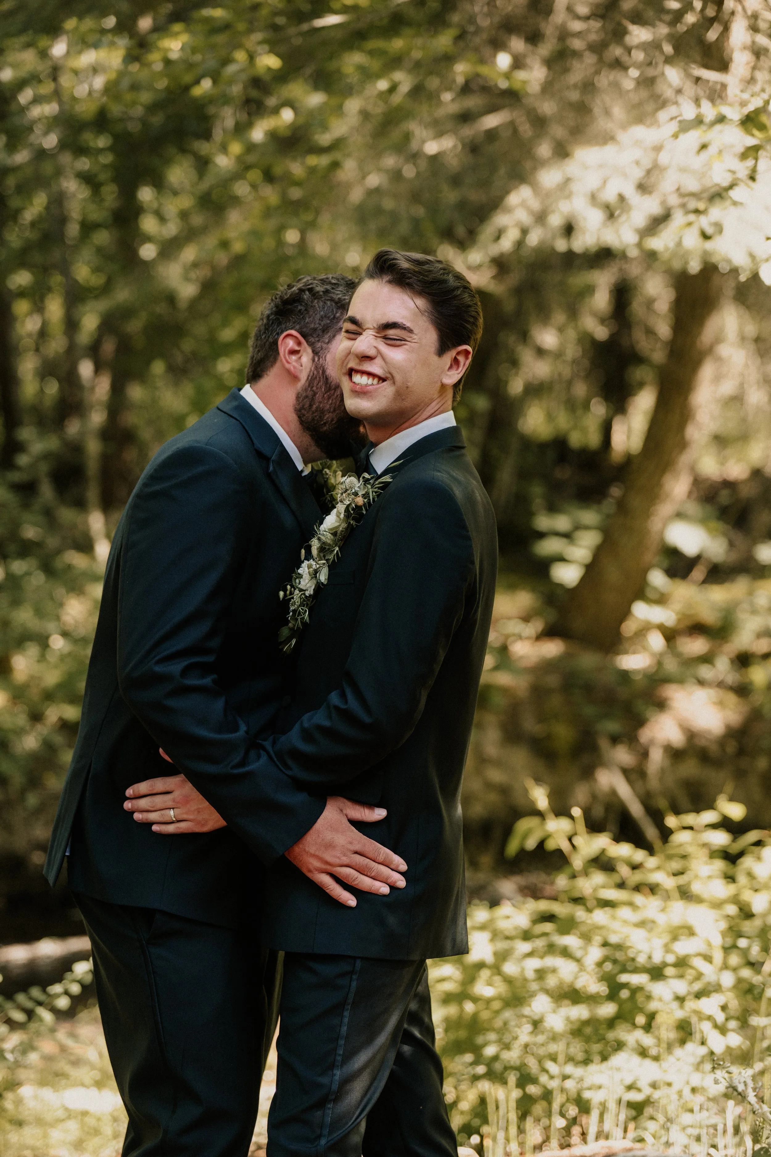 Two men in suits hugging and smiling in a lush forest during a wedding or special event.