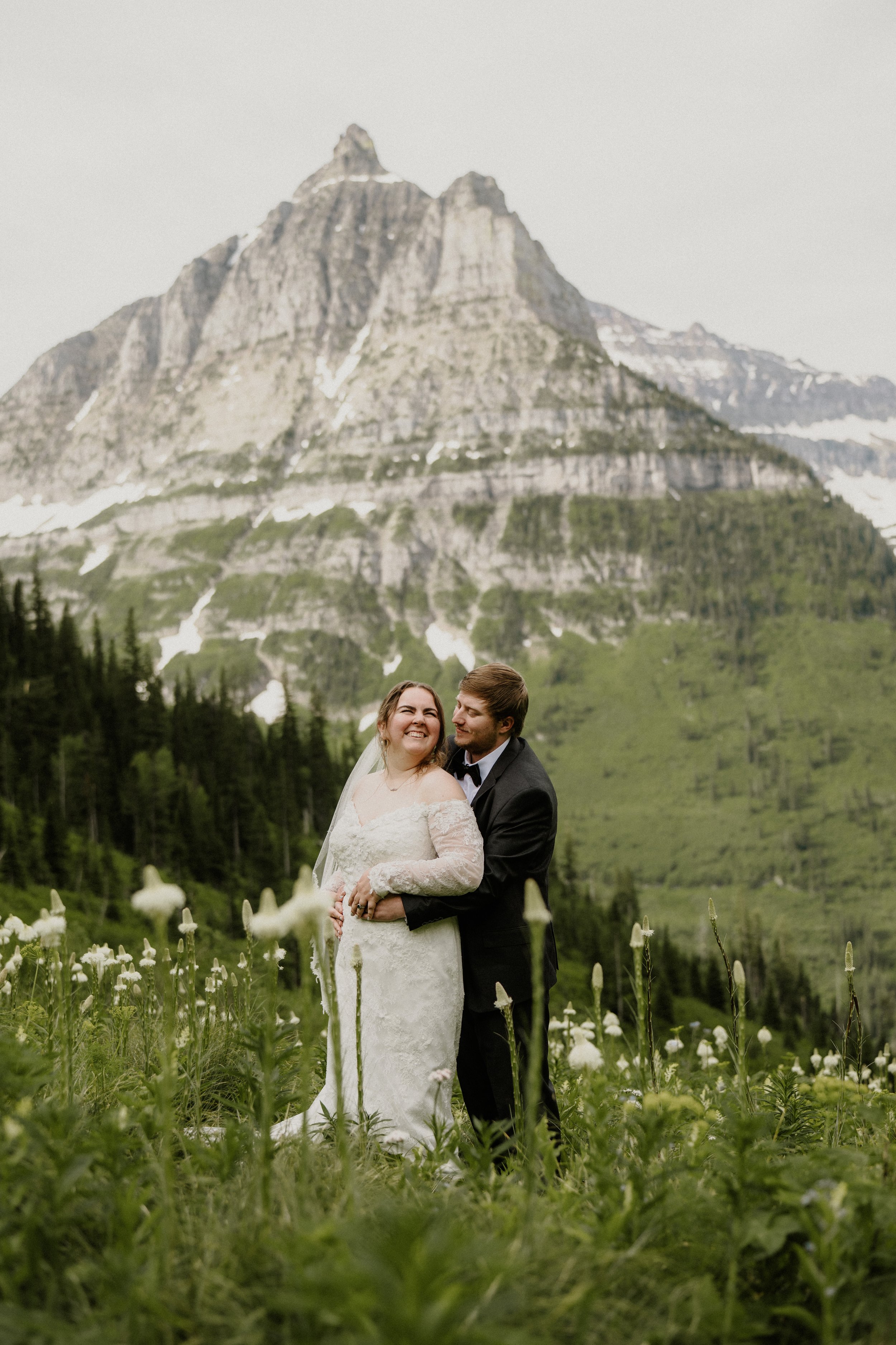 A newlywed couple standing in a field of white flowers with a mountain range in the background. The bride is smiling and wearing a white lace wedding dress, and the groom is embracing her, smiling at her, and wearing a black tuxedo. The scene is outd