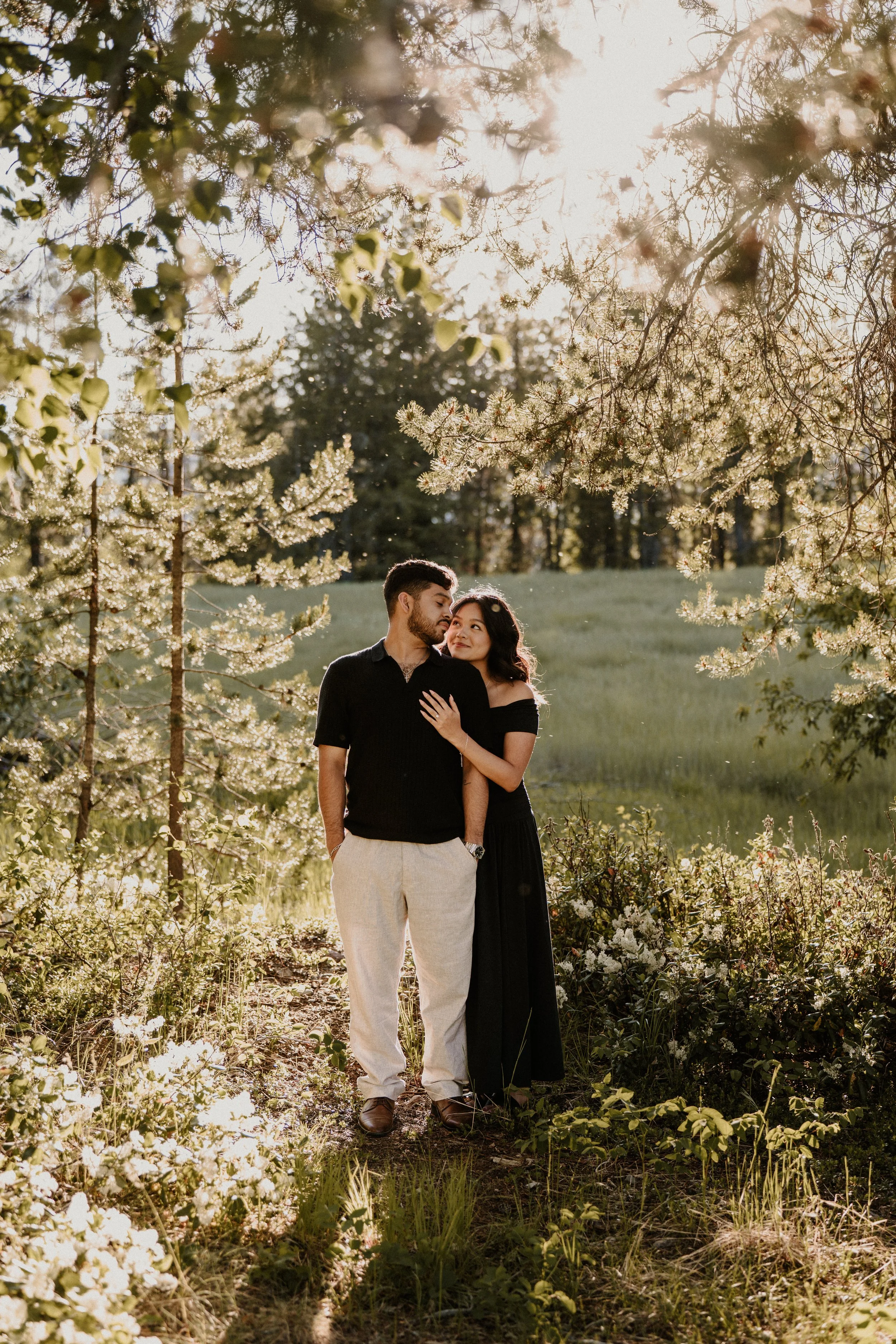 Couple standing close together outdoors during sunset surrounded by trees and wildflowers in Glacier National Park
