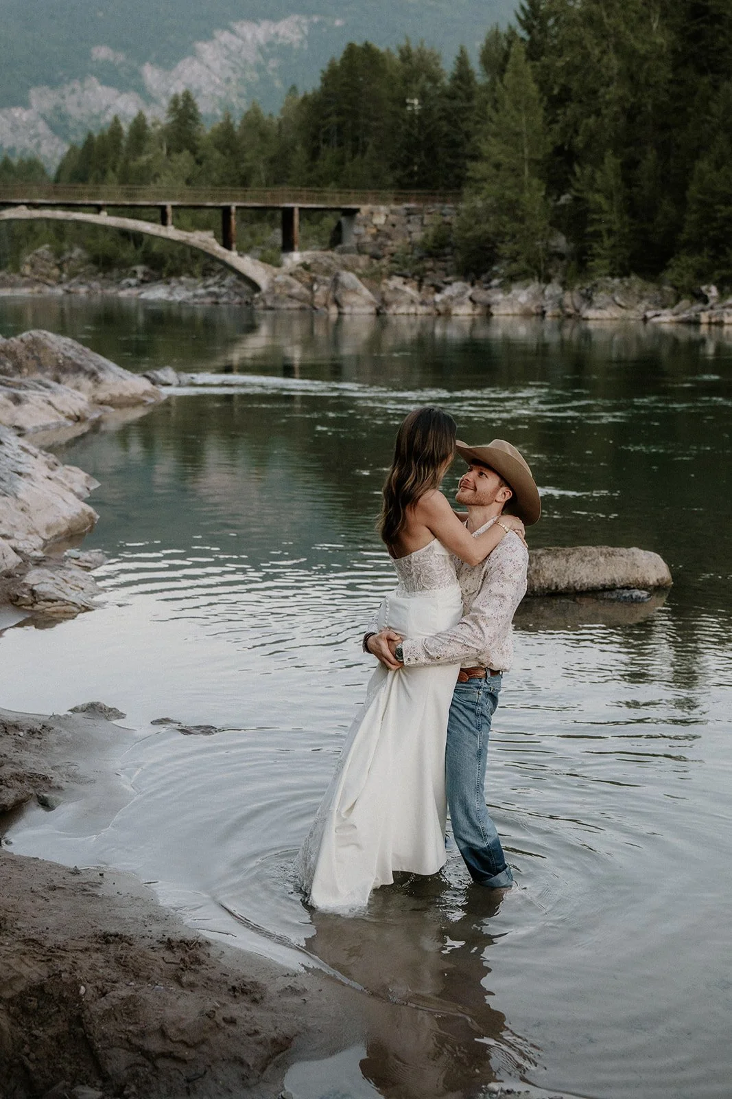A couple standing in a river with a scenic mountain and forest background, embracing, with the woman in a white dress and the man in a cowboy hat and western attire.