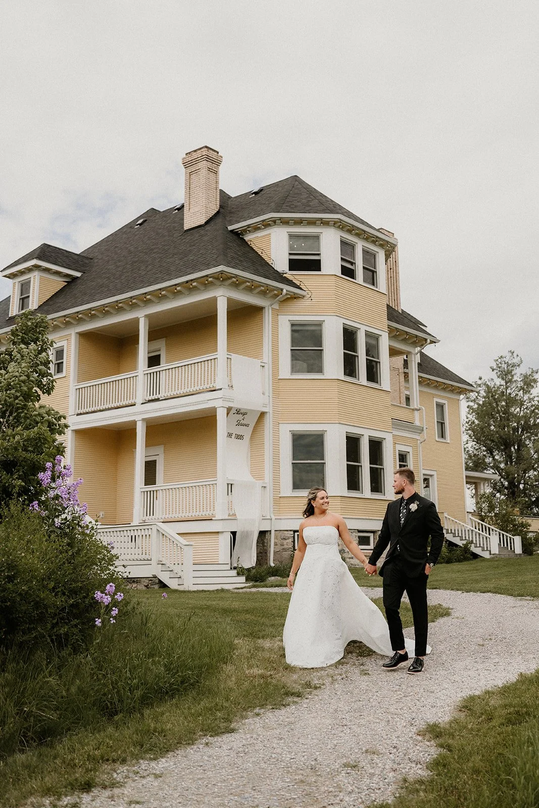 A bride and groom holding hands walk on a gravel path in front of a large yellow Victorian-style house on a cloudy day.