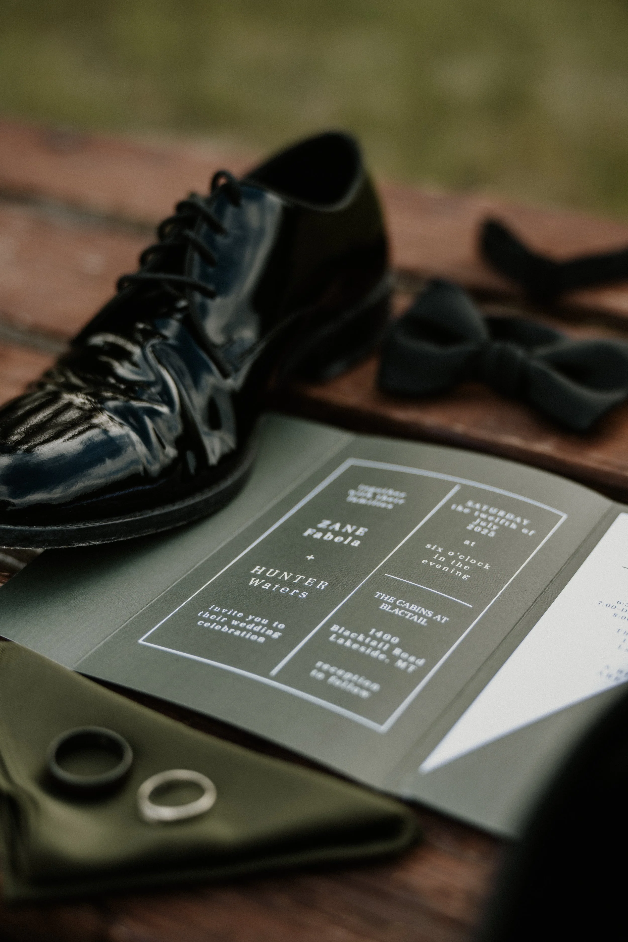 A pair of shiny black women's oxford shoes, a black bow tie, a wedding invitation, a green fabric pouch with rings, and a wooden surface.