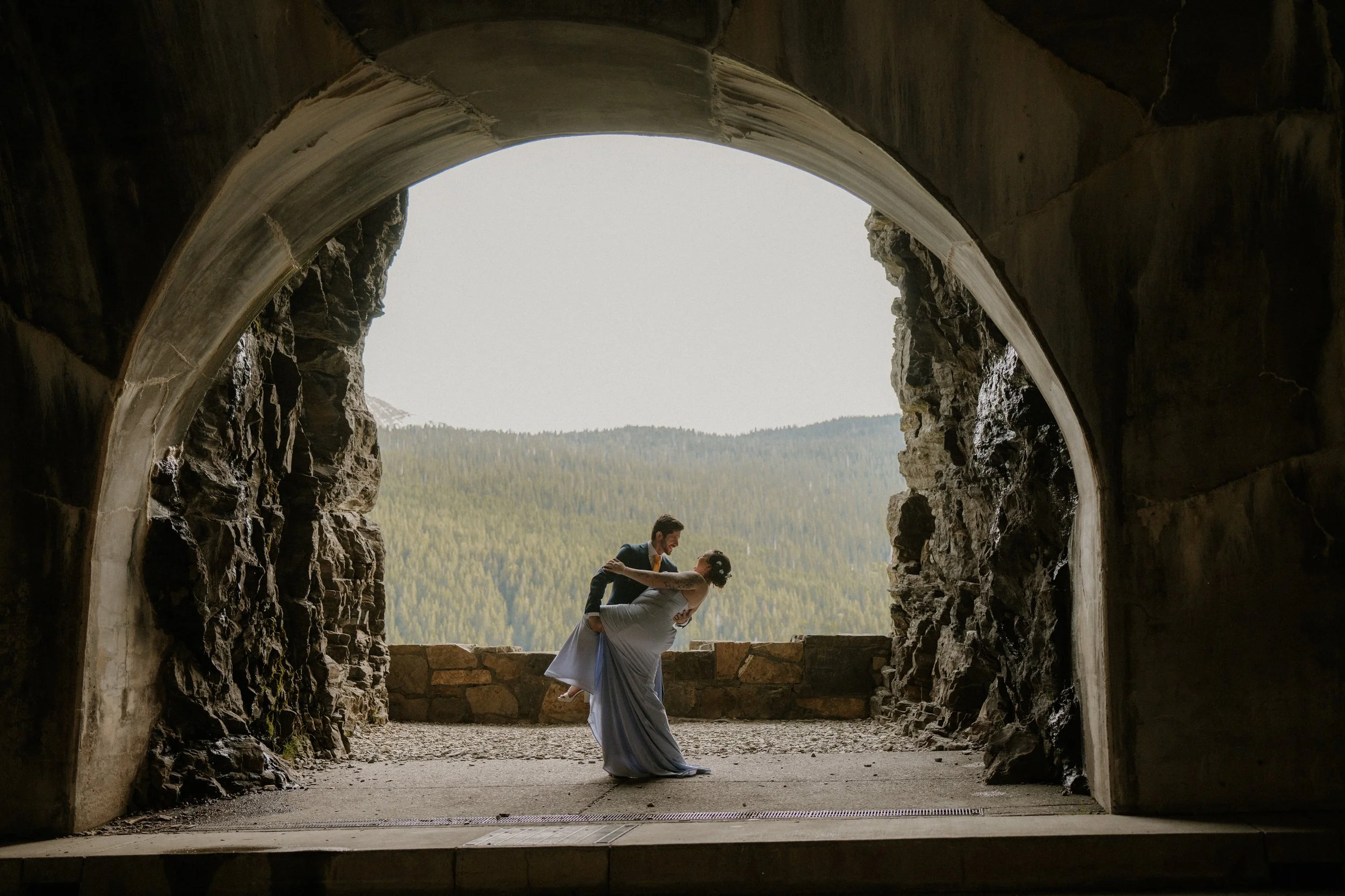 A couple dancing, with the man in a suit and the woman in a white dress, dancing on a stone ledge inside a tunnel with a view of mountains and forest outside.