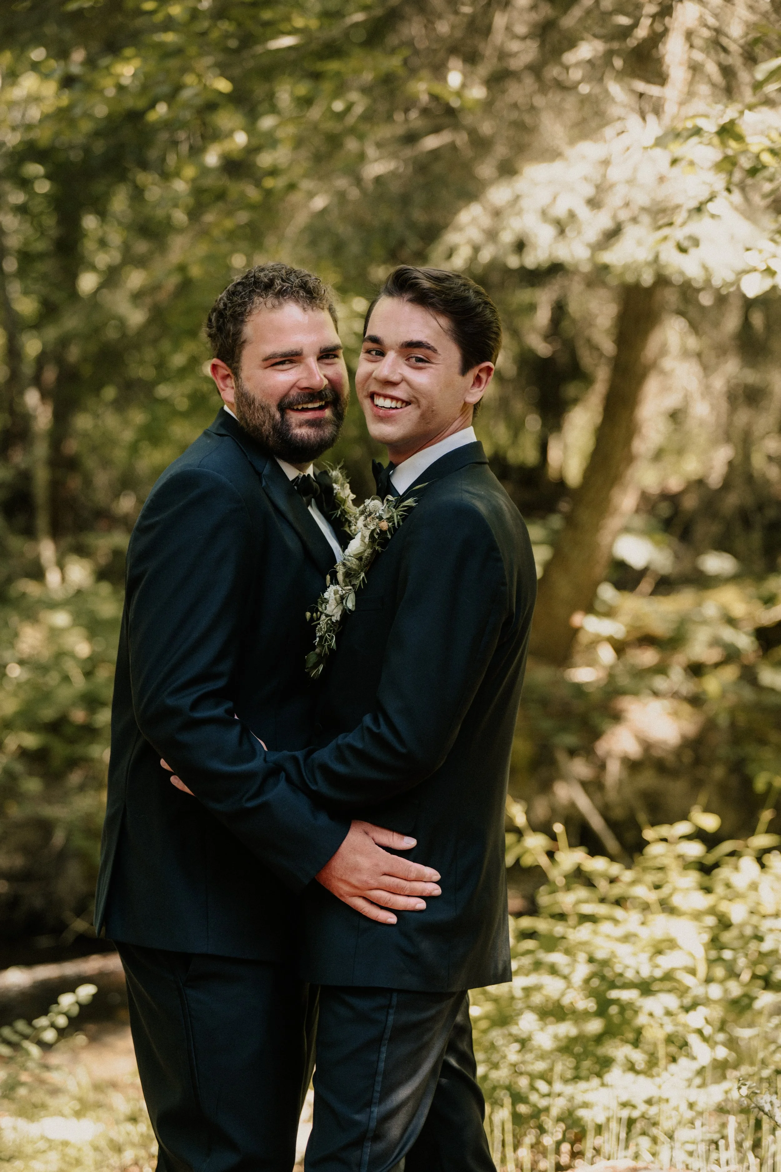 Two men in tuxedos smiling and embracing in an outdoor wooded setting.
