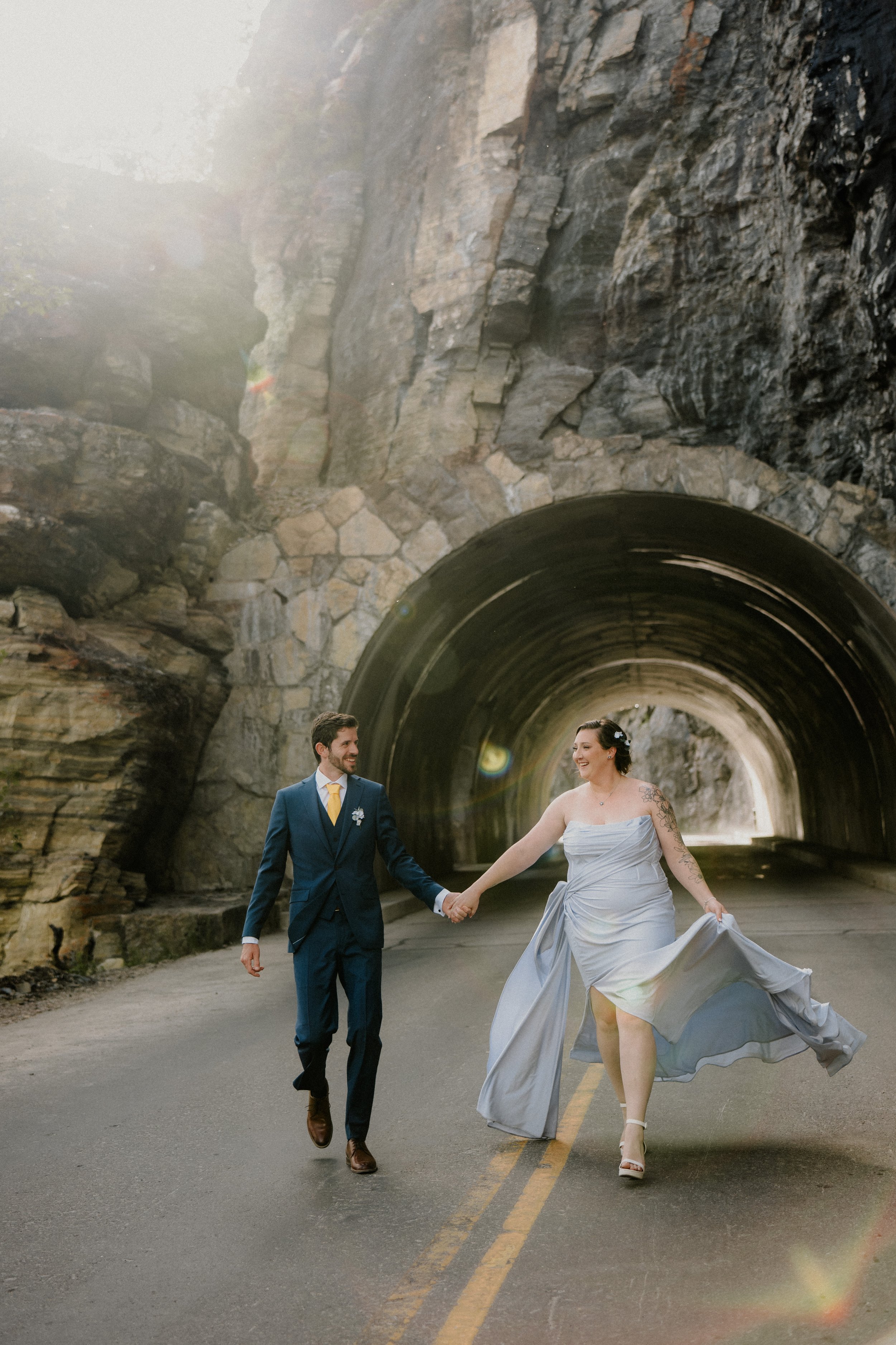 A bride and groom holding hands and walking through a tunnel under a rocky hillside. The groom is wearing a blue suit with a yellow tie, and the bride is in a strapless white wedding dress, lifting her dress slightly with her hand. They are smiling a
