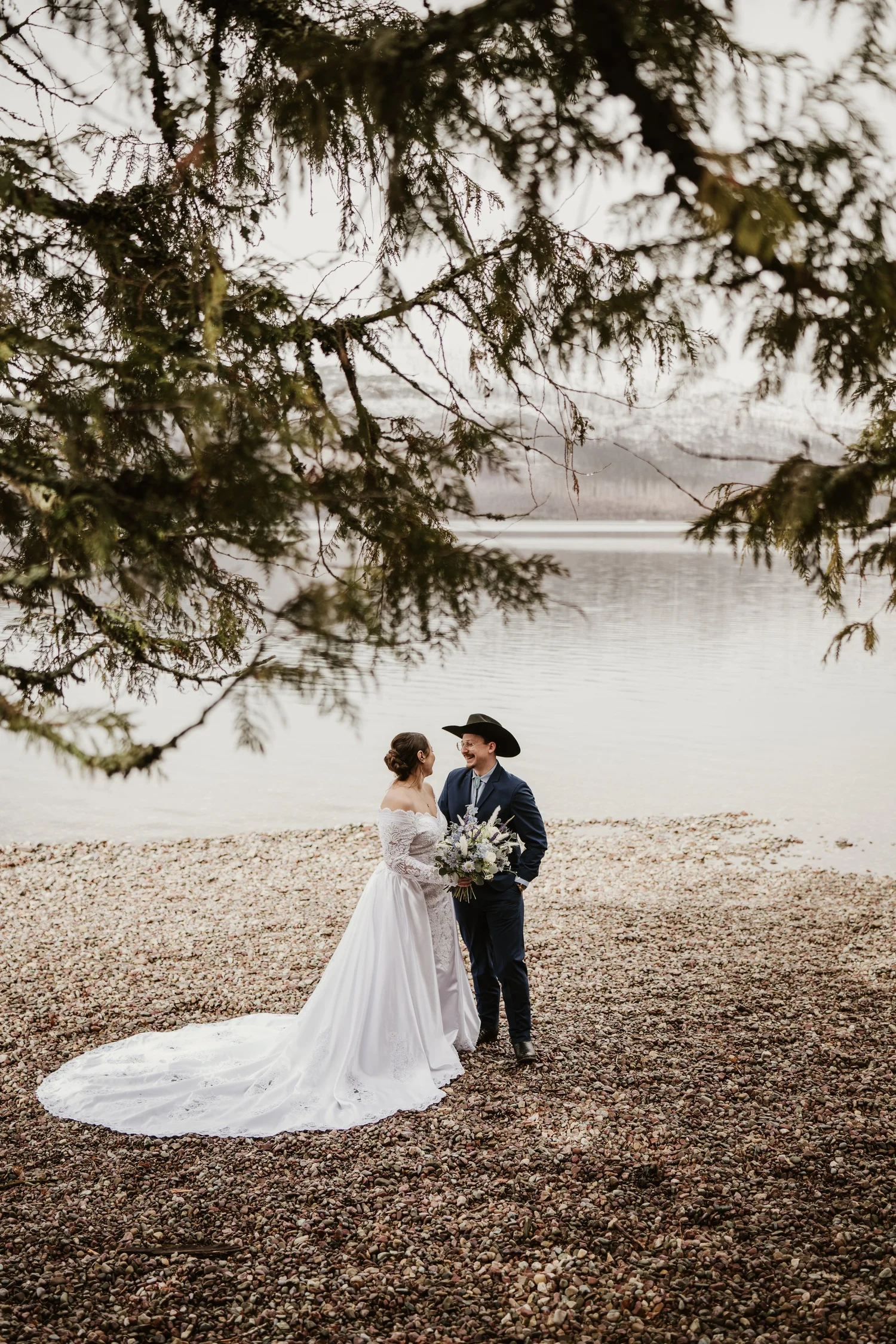 Bride and groom on a pebbled lakeshore framed by branches, mountains behind