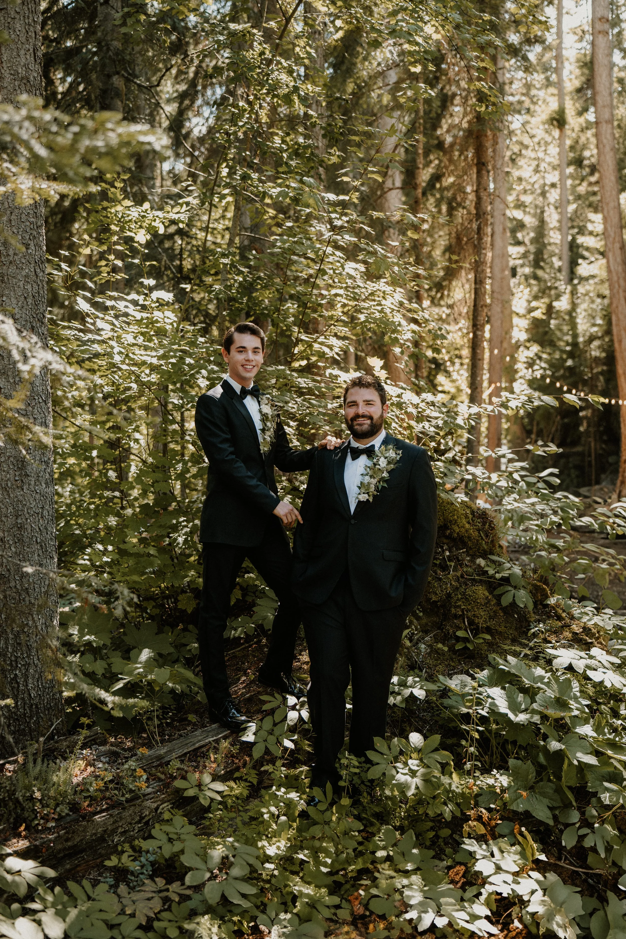 Two men in tuxedos with floral boutonnieres, standing in a forest with tall trees and green foliage, smiling at the camera, during a wedding or formal event.