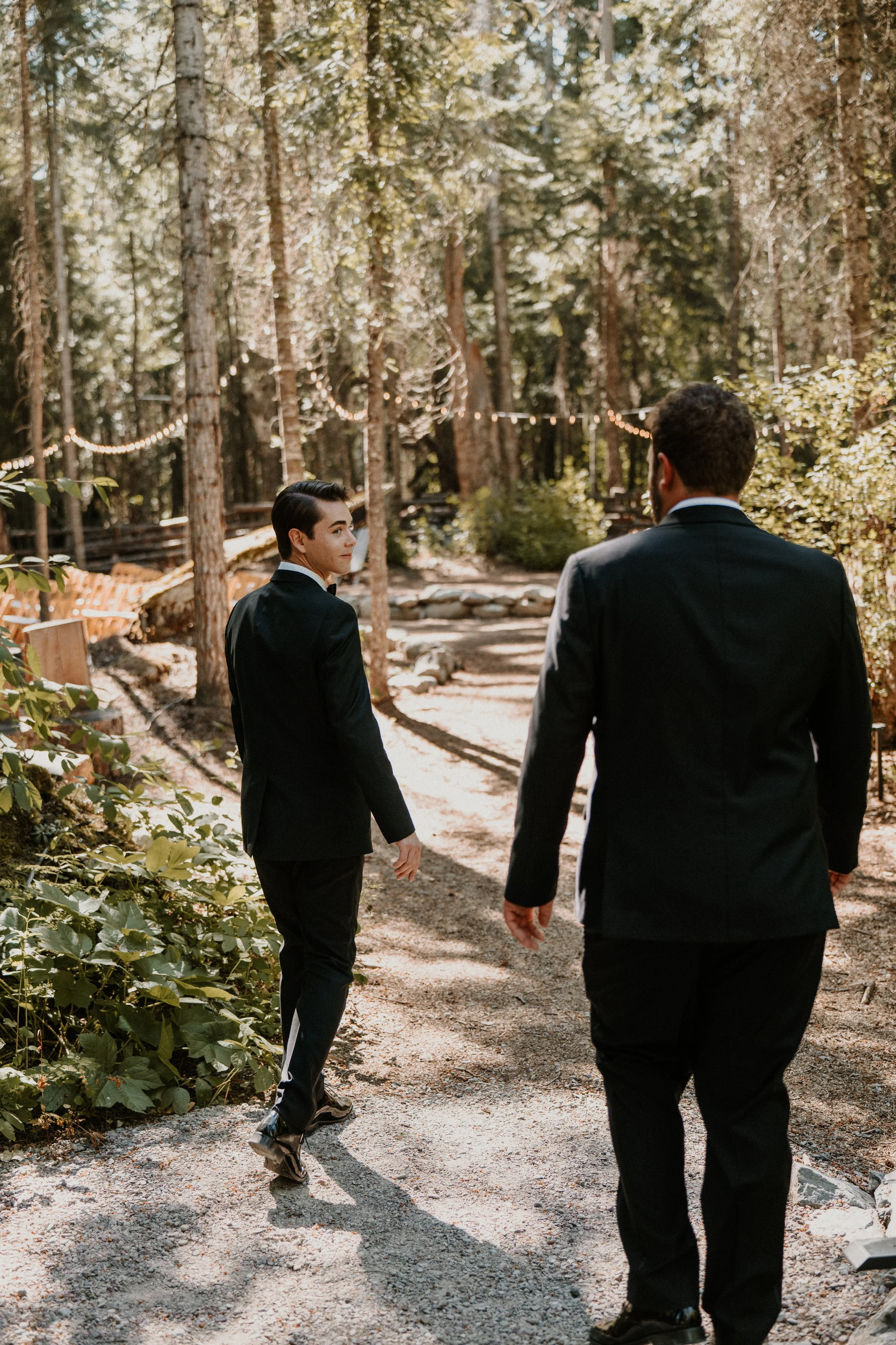 Two men in formal suits walking along a dirt path in a wooded area, with one looking back over their shoulder. The scene is illuminated by natural sunlight and string lights hanging among the trees.
