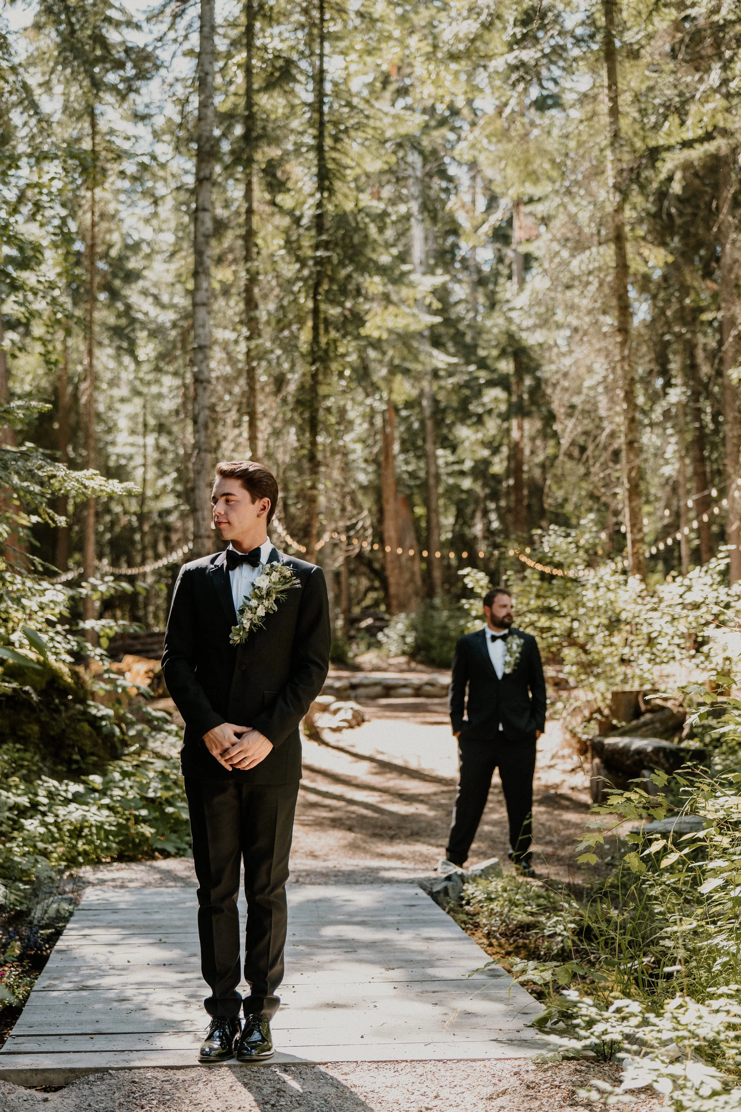 Two men in tuxedos standing on a forest path, with greenery and string lights in the background.