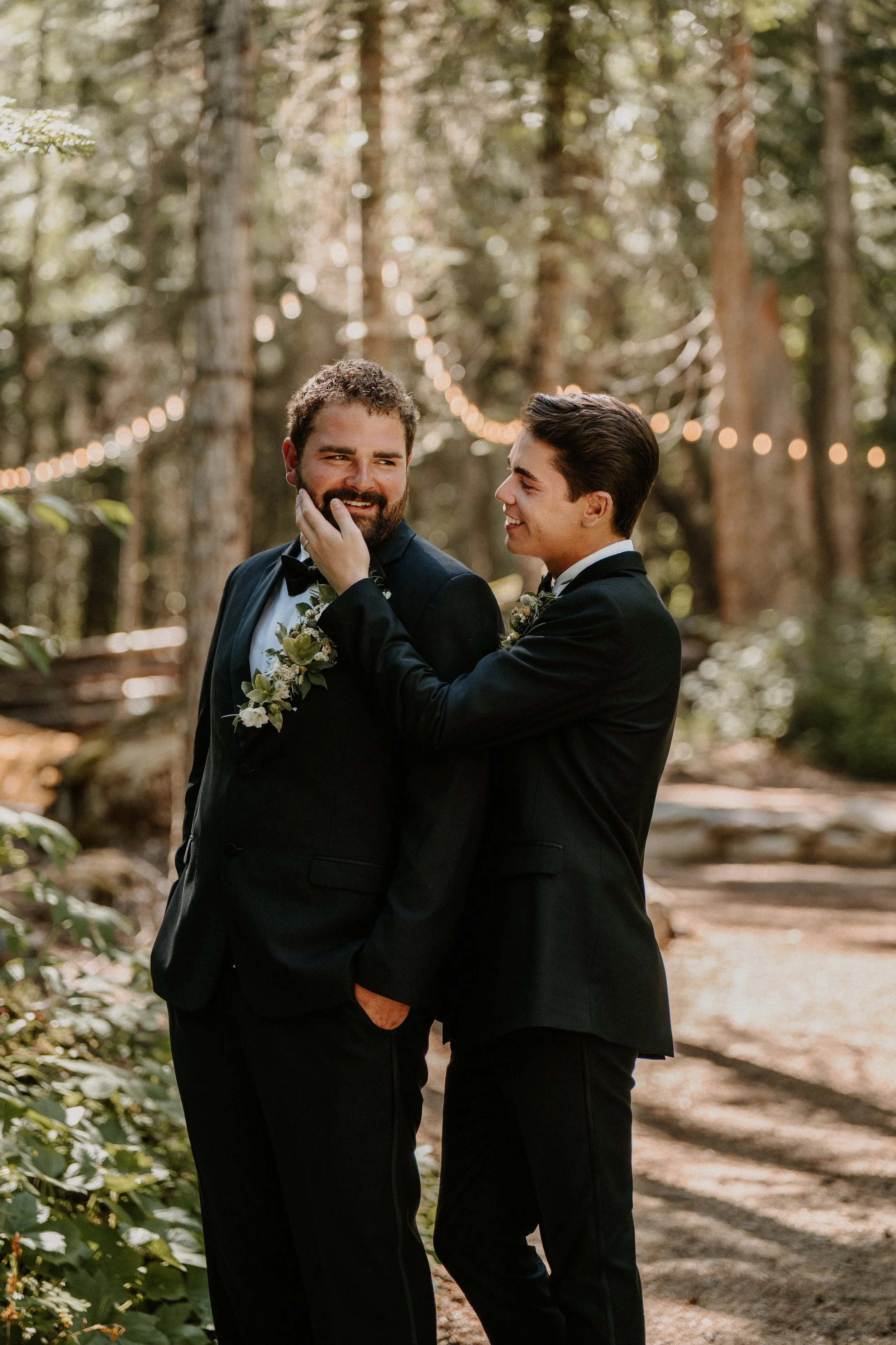 Two men in black tuxedos sharing a joyful moment in a wooded outdoor setting, with string lights hanging in the background.