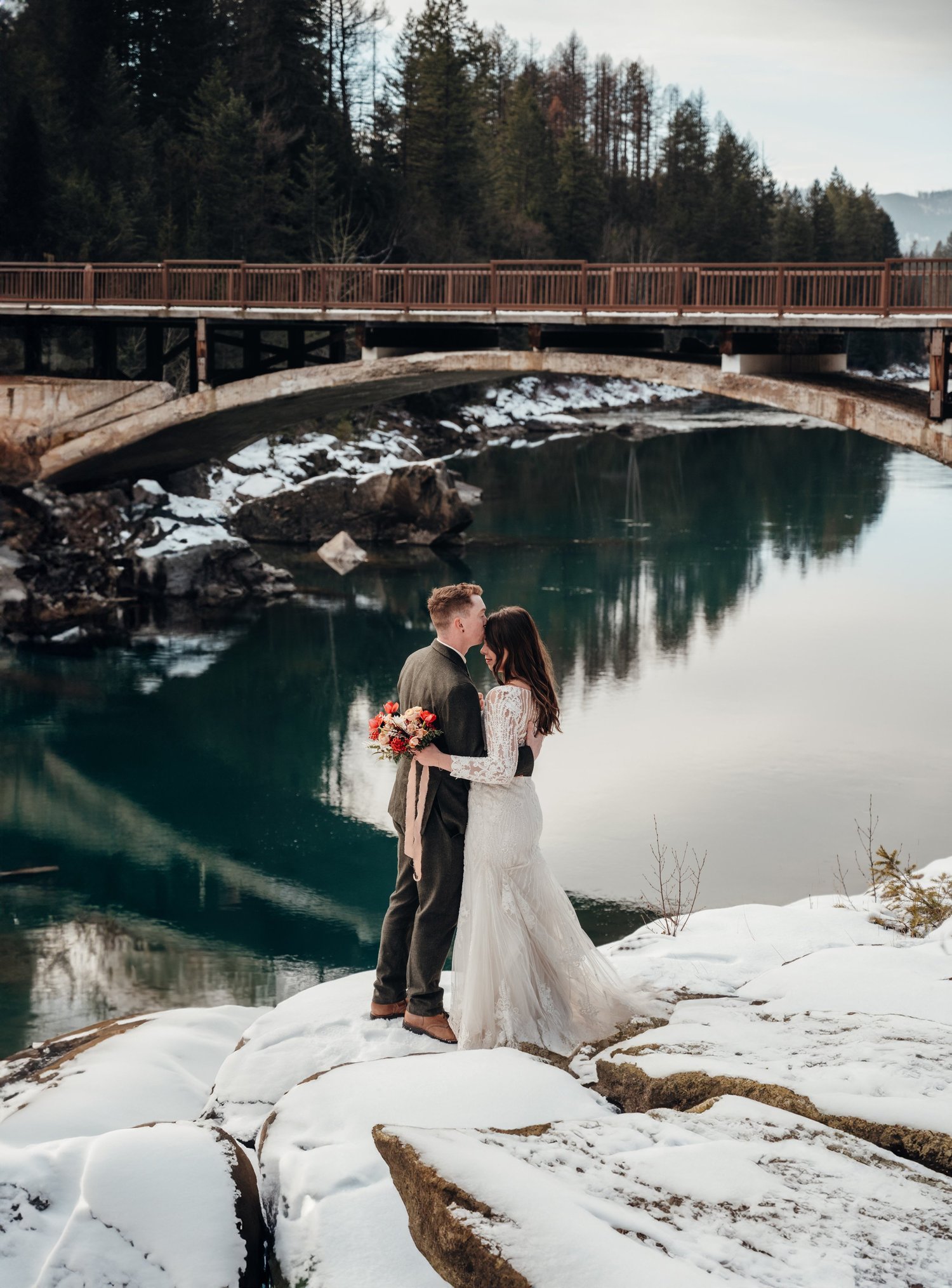 Couple in wedding attire standing on snow-covered rocks by McDonald Creek near the bridge