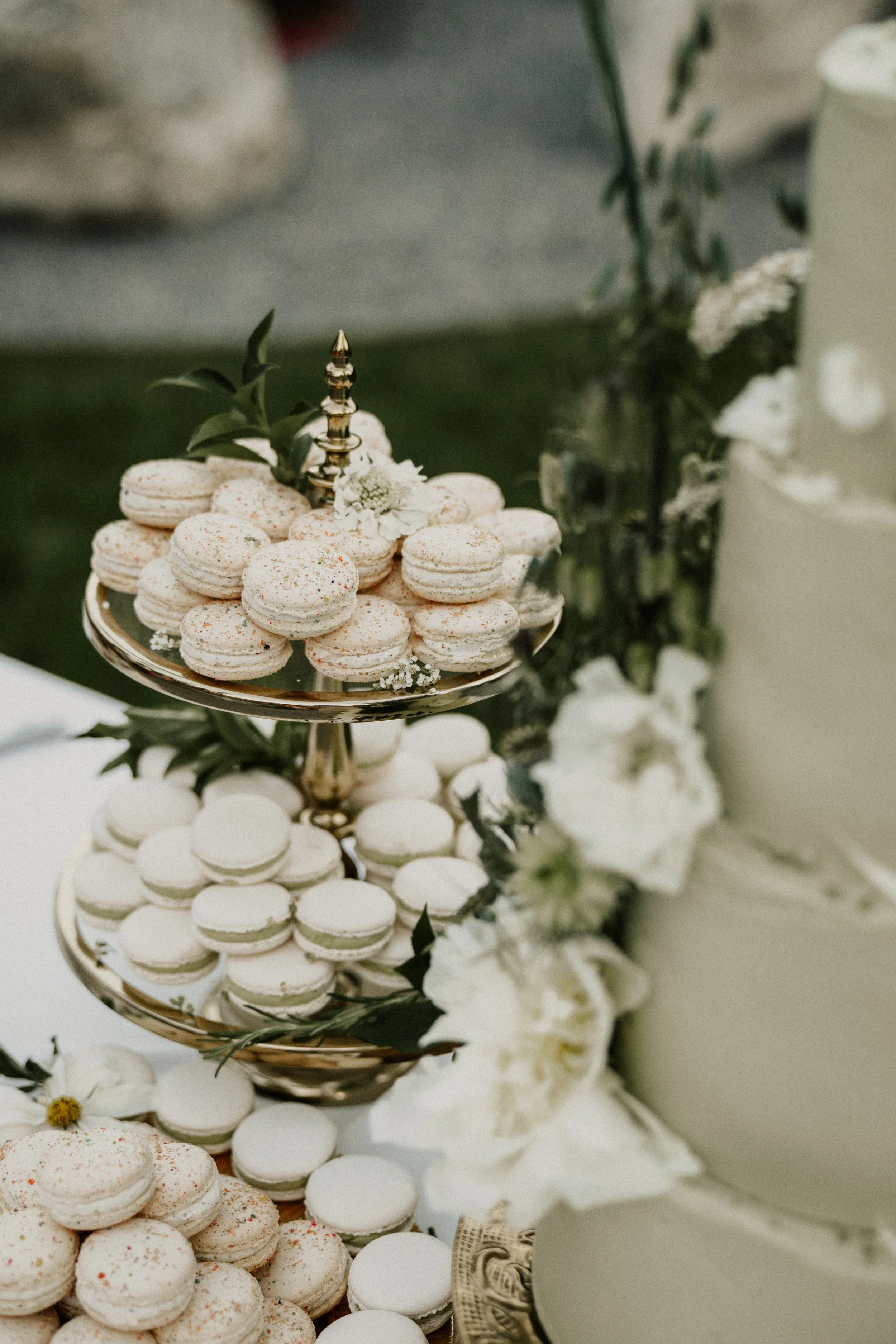 A dessert table with a tiered platter holding white macarons decorated with sprinkles and floral accents, next to a three-tiered white wedding cake adorned with white flowers.