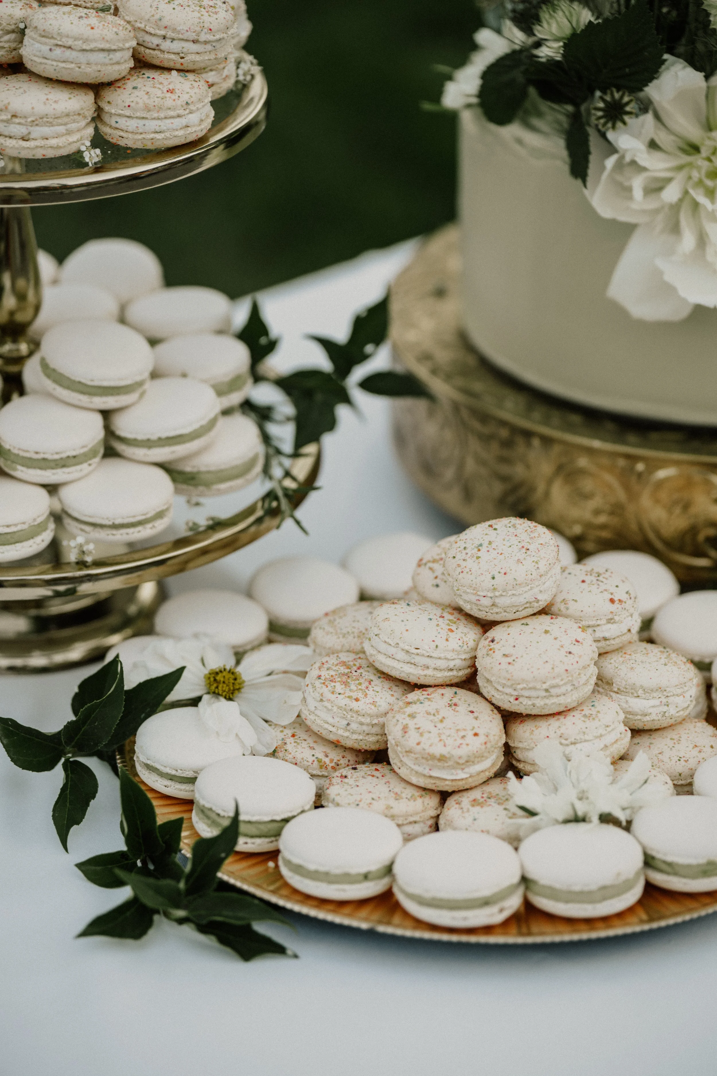 Assorted white macarons with some sprinkled with colorful sugar, arranged on gold and wooden trays, decorated with green leaves and white flowers, with a white cake with flowers in the background.