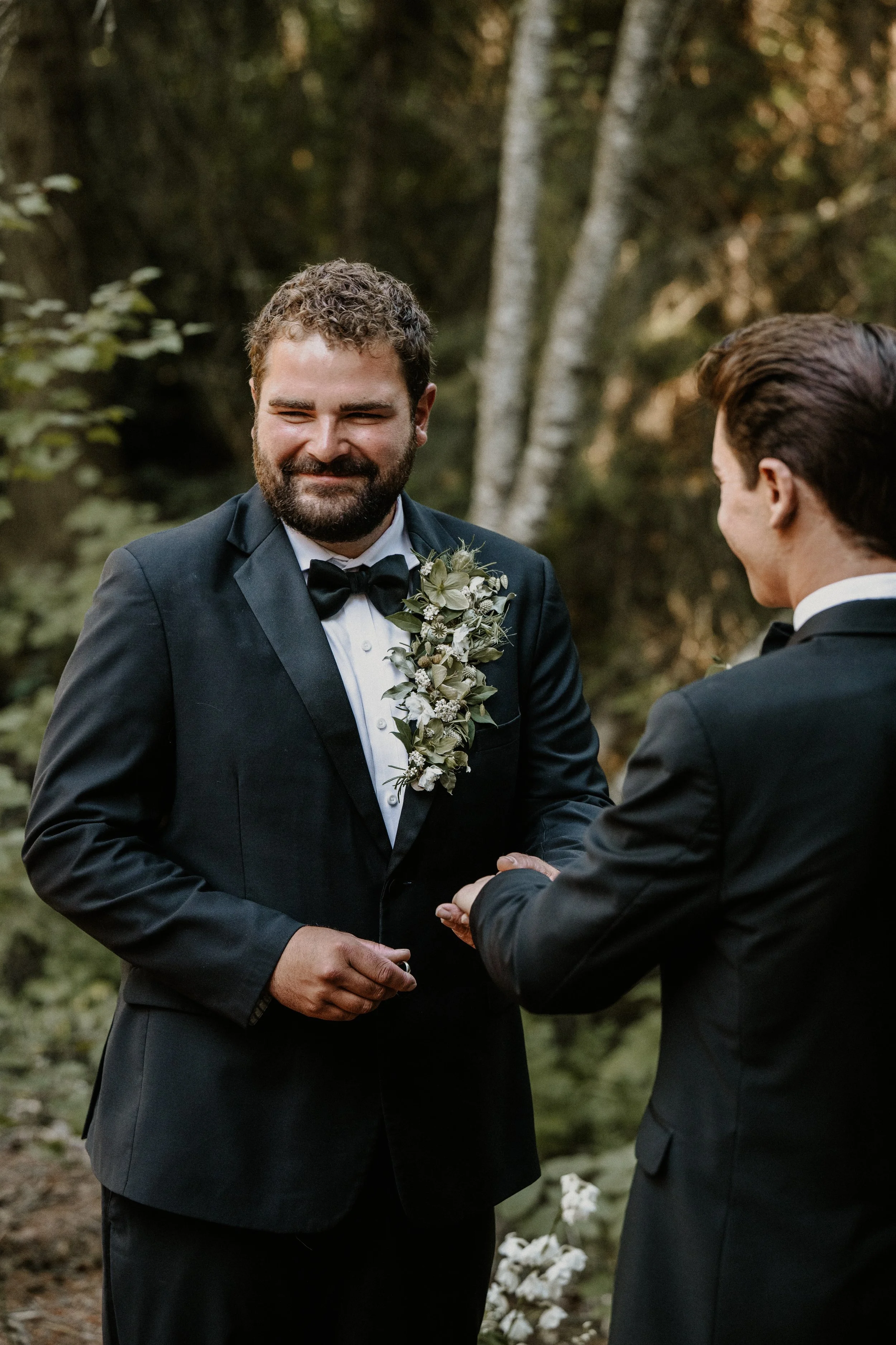 Two men in suits exchanging rings in a forest setting during a wedding ceremony.