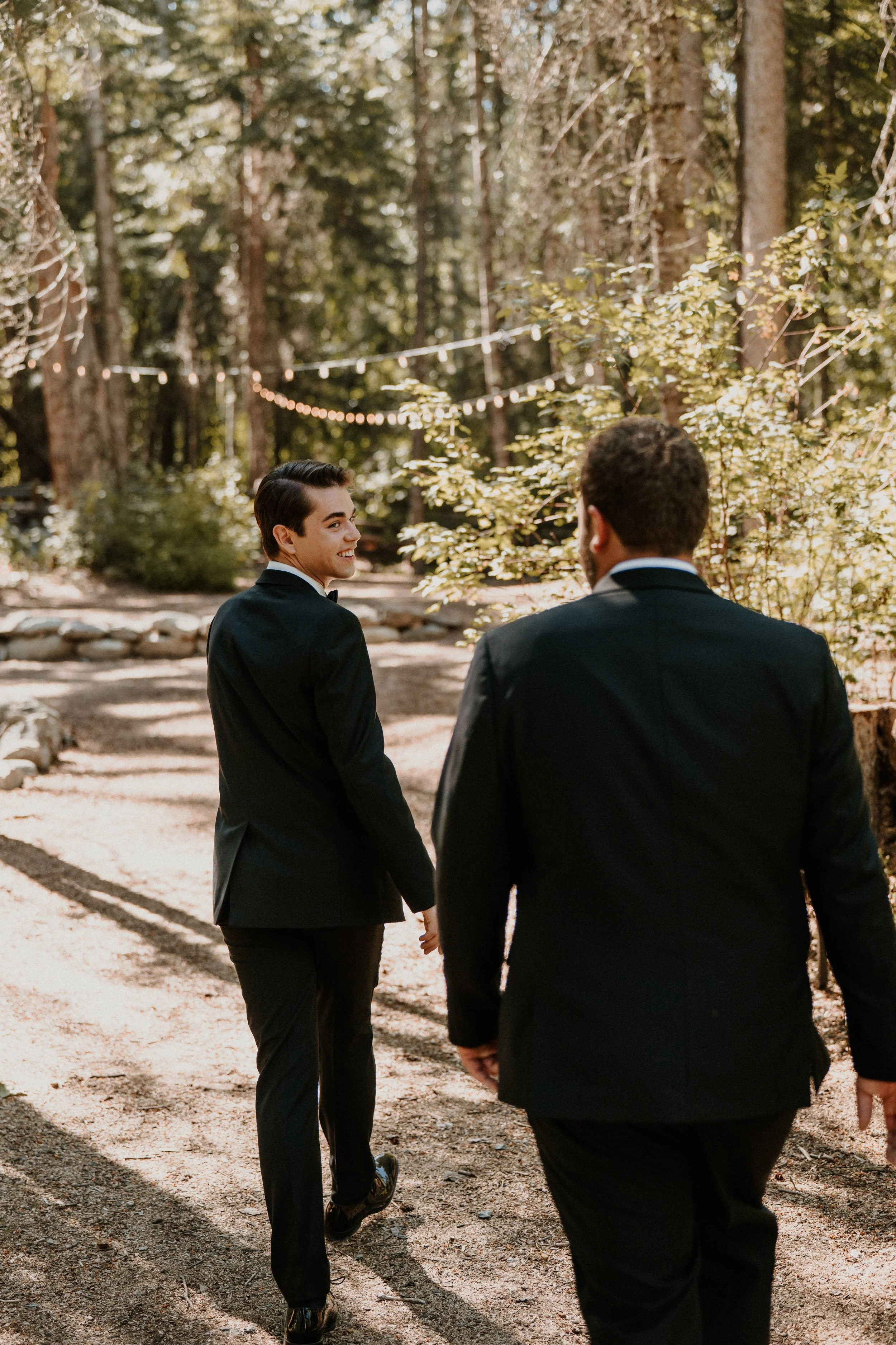 Two men in black suits walking hand-in-hand through a wooded area with string lights overhead, one smiling at the other.