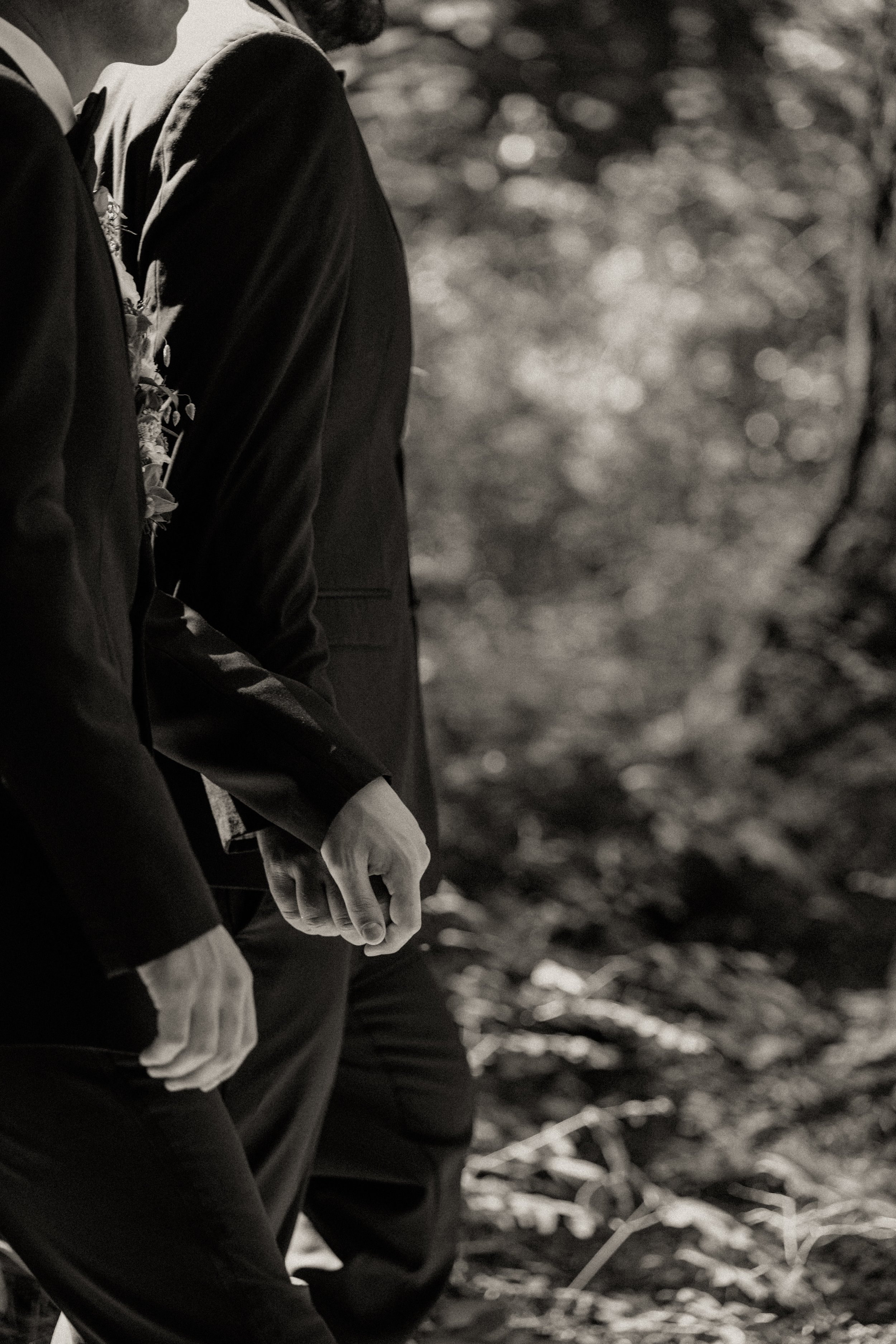 Two men wearing suits holding hands in an outdoor wooded setting.