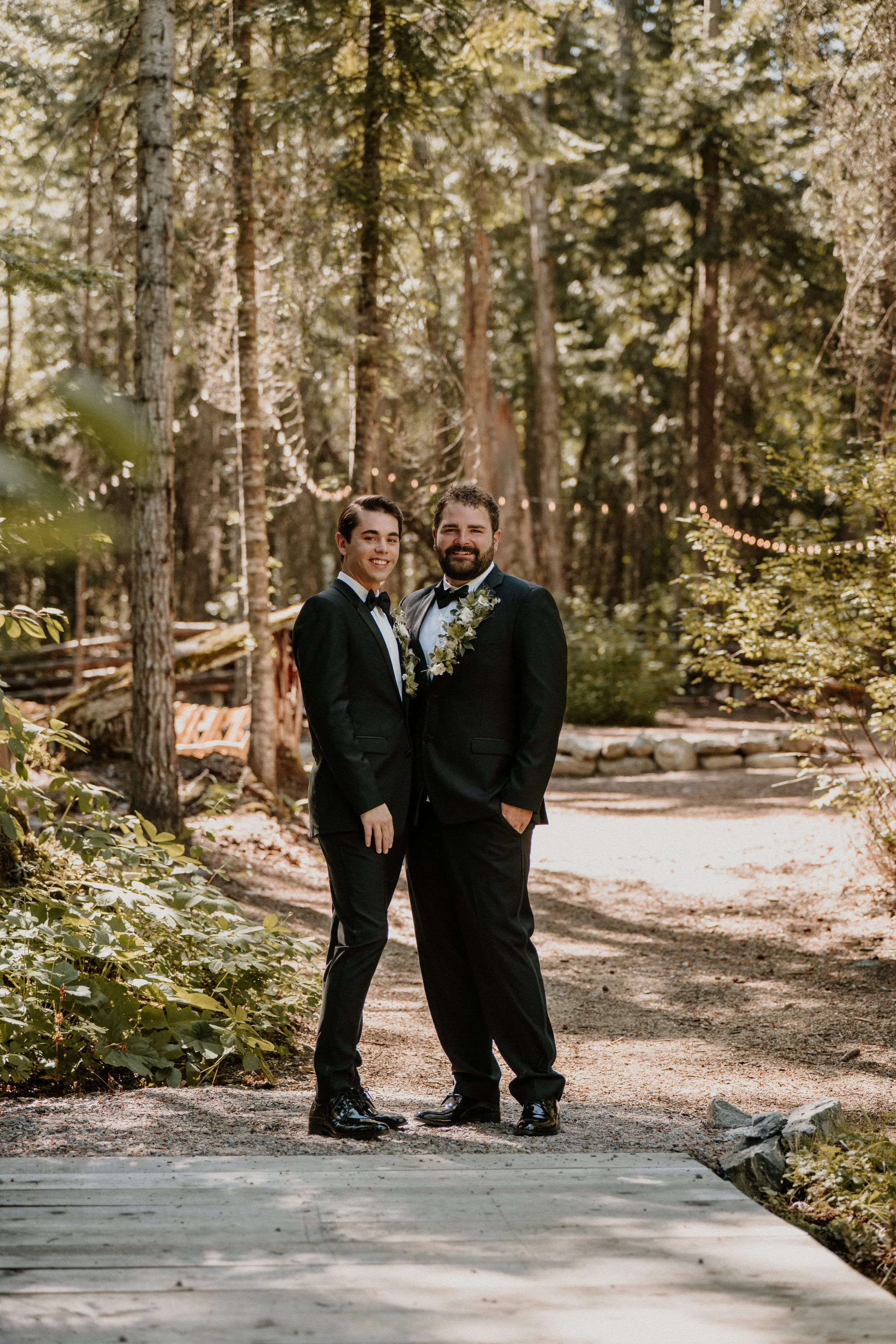 Two men in tuxedos standing close together outdoors in a wooded area, smiling, with string lights in the background.