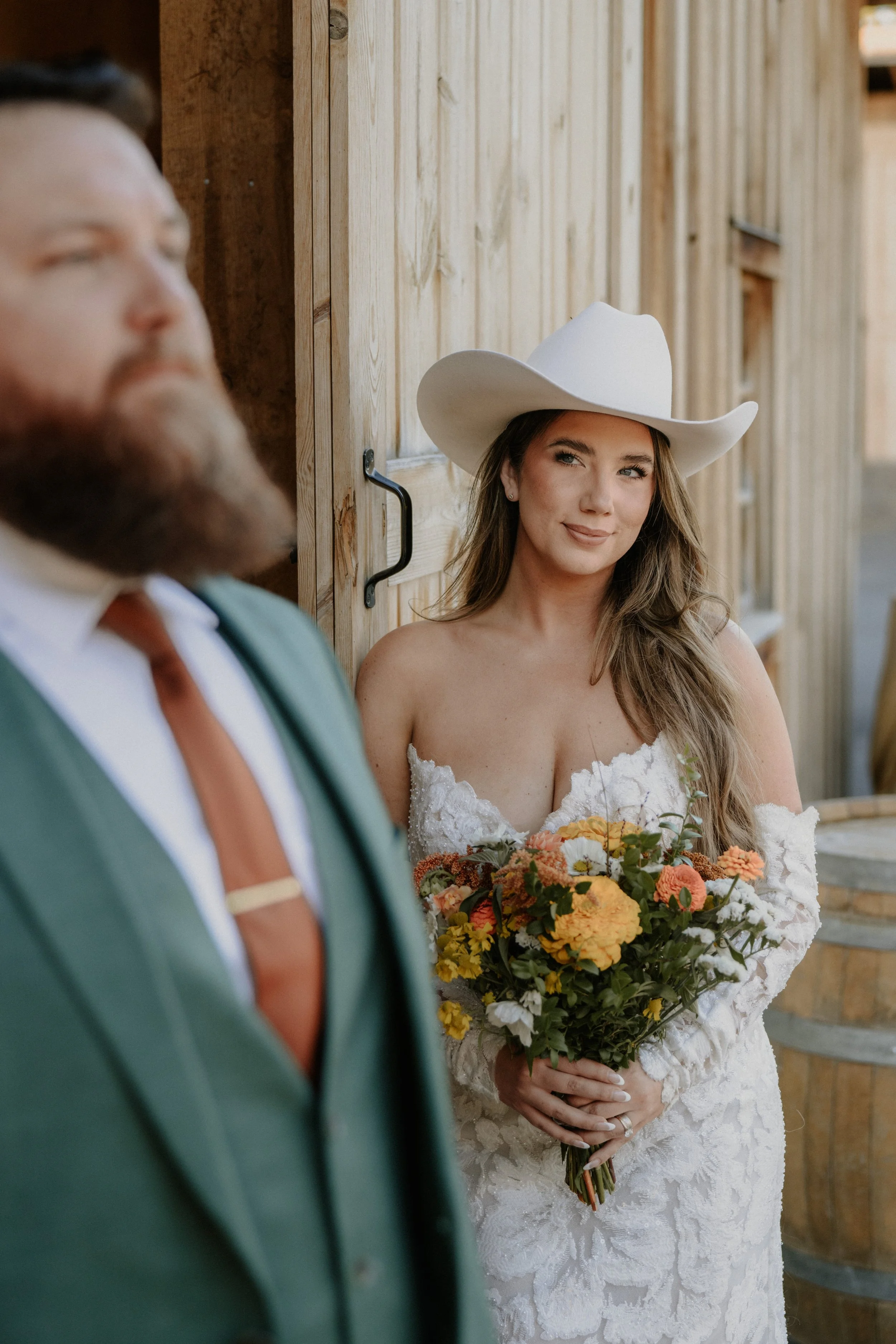 A bride in a white lace dress and cowboy hat holding a bouquet of colorful flowers, smiling softly, standing beside her groom in a suit with a bow tie, at a wooden rustic setting.