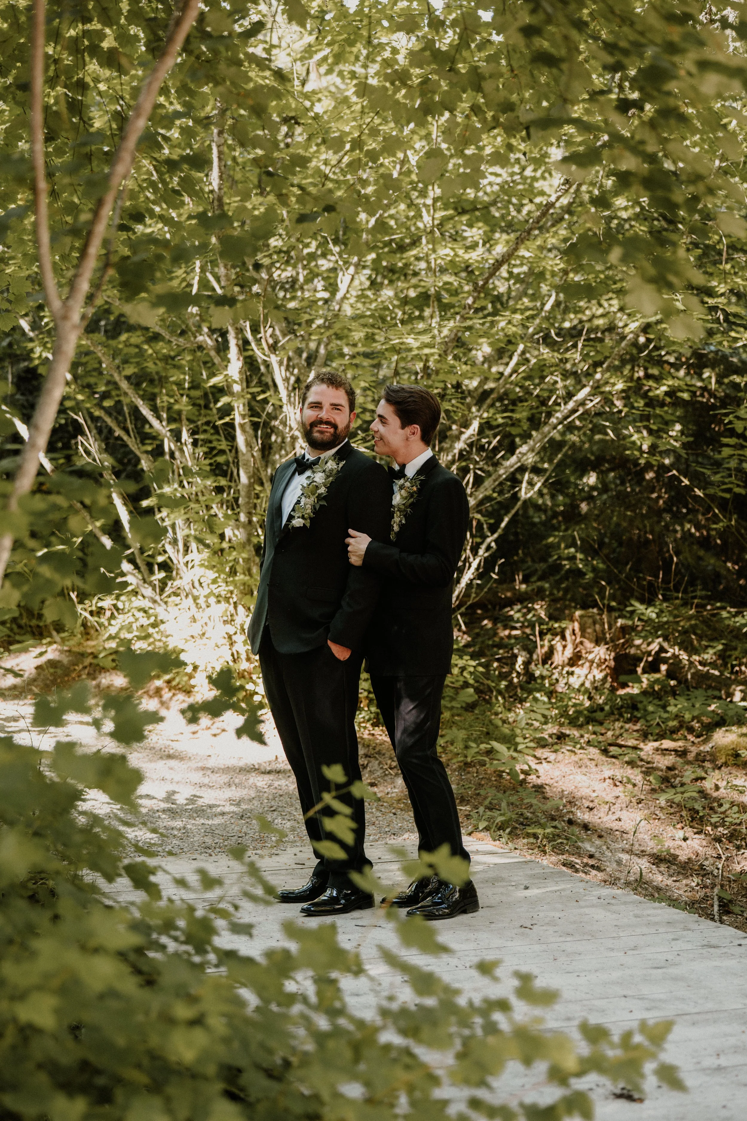 Two men in tuxedos with floral boutonnieres standing on a wooden path in a wooded area, smiling and embracing each other.