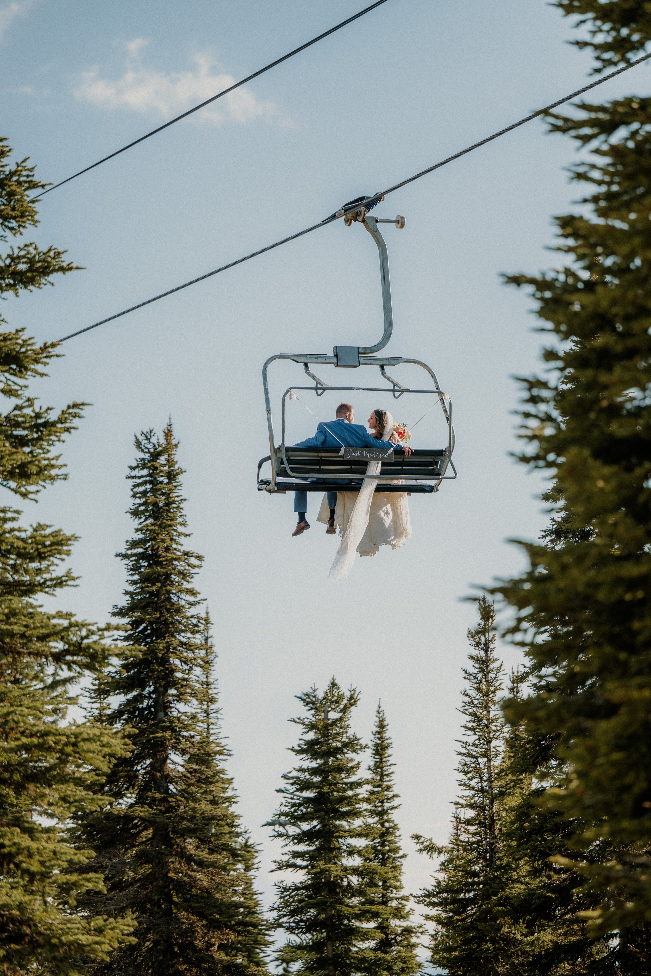 A couple is sitting on a ski lift or chairlift, surrounded by tall pine trees, in an outdoor setting during daylight. The couple appears to be embracing or talking, and the sign on the lift reads 'Just Married.'